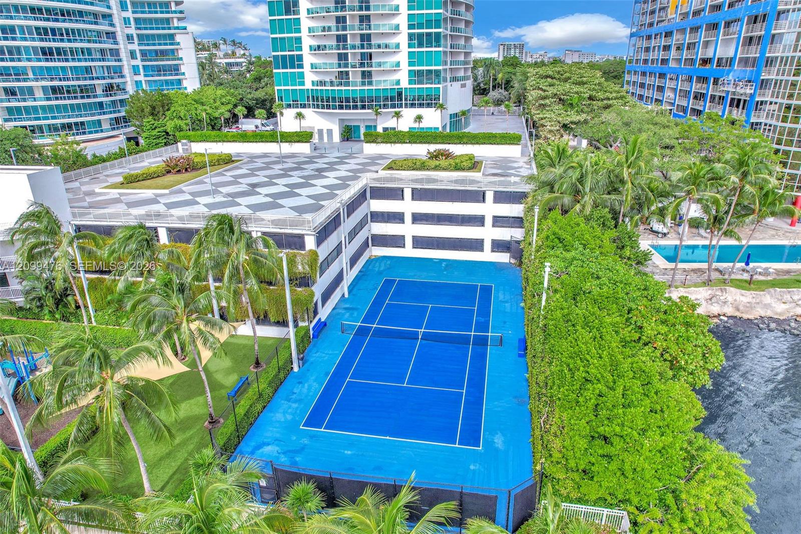 Tennis court with blue hard-surface surfacing, enclosed by perimeter fencing, surrounded by mature palm trees, landscaped grounds, and a swimming pool