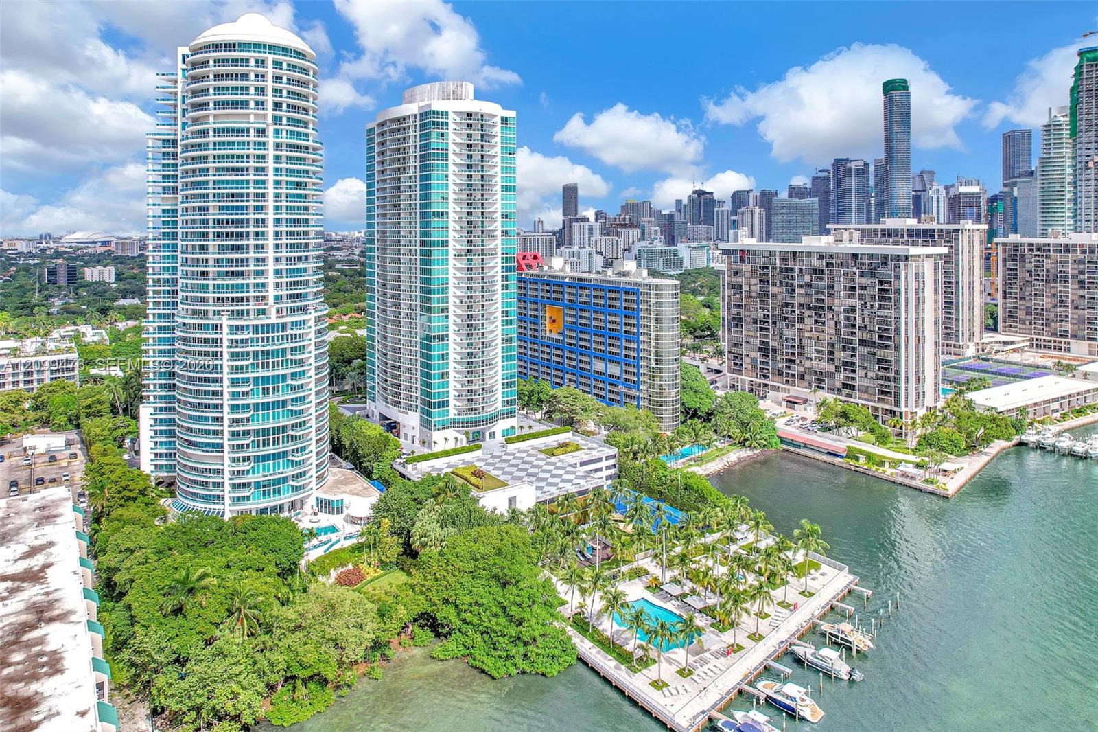 High-rise residential towers featuring expansive glass-paneled balconies, a ground-level swimming pool, manicured grounds with mature palm trees, and a private marina docking area