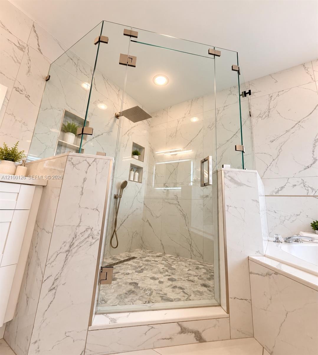 Walk-in shower featuring floor-to-ceiling stone-style tile, frameless glass enclosure, rainfall showerhead, handheld sprayer, and recessed wall shelving