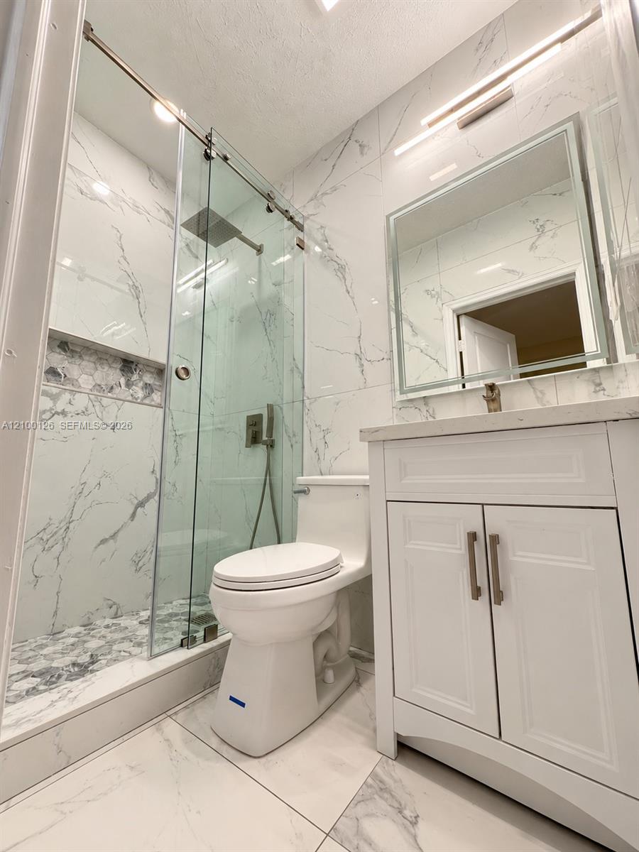 Full-height stone-finish wall tiling, glass-enclosed shower with a rainfall showerhead, white vanity with stone-finish countertop, backlit mirror, and integrated shower wall niche