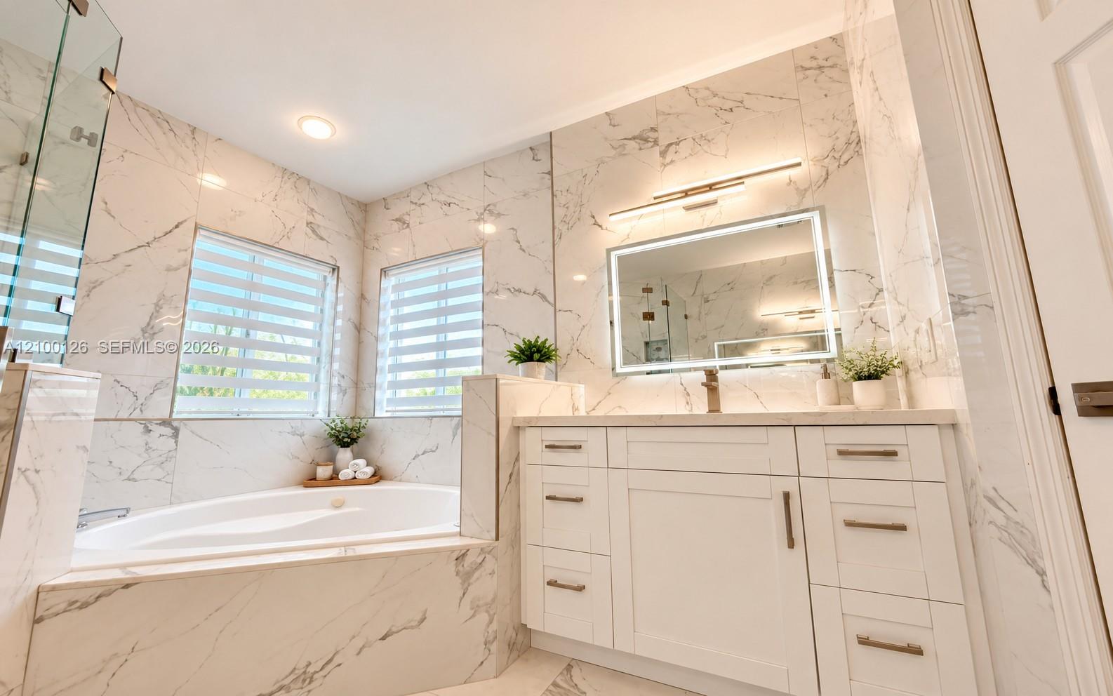 Floor-to-ceiling veined stone tile, white shaker-style vanity with brass-finish hardware, built-in soaking tub, backlit vanity mirror, and glass-enclosed shower