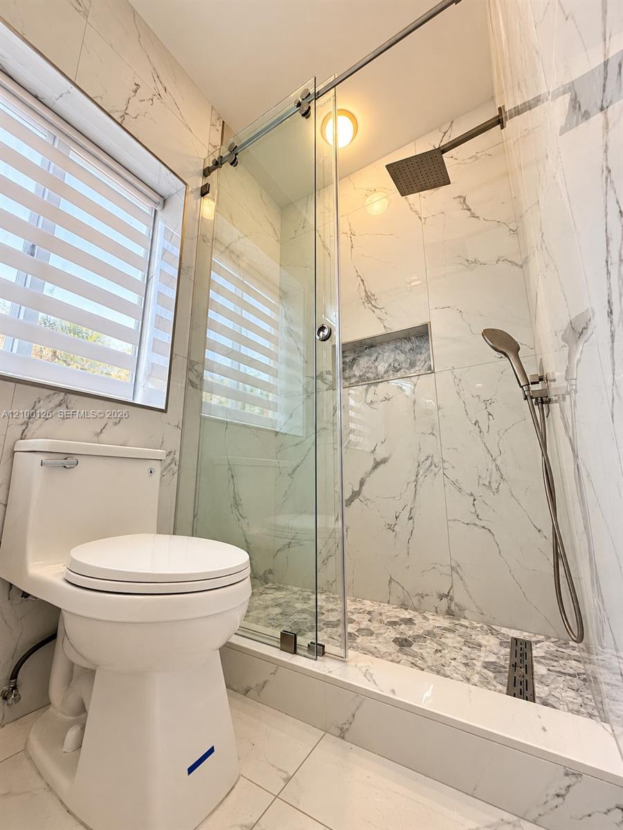 Walk-in shower featuring sliding glass doors, ceiling-mounted rainfall showerhead, handheld sprayer, integrated wall niche, and natural stone-finish floor tile