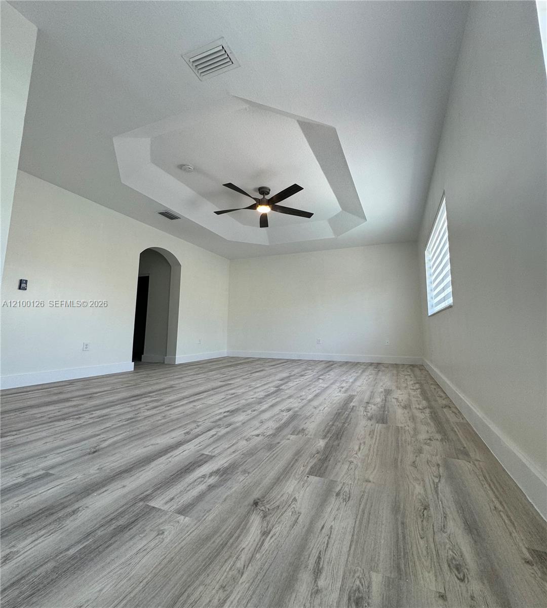 Tray ceiling with a central multi-blade ceiling fan, wood-finish flooring, arched entryway, and dual-layered window shades