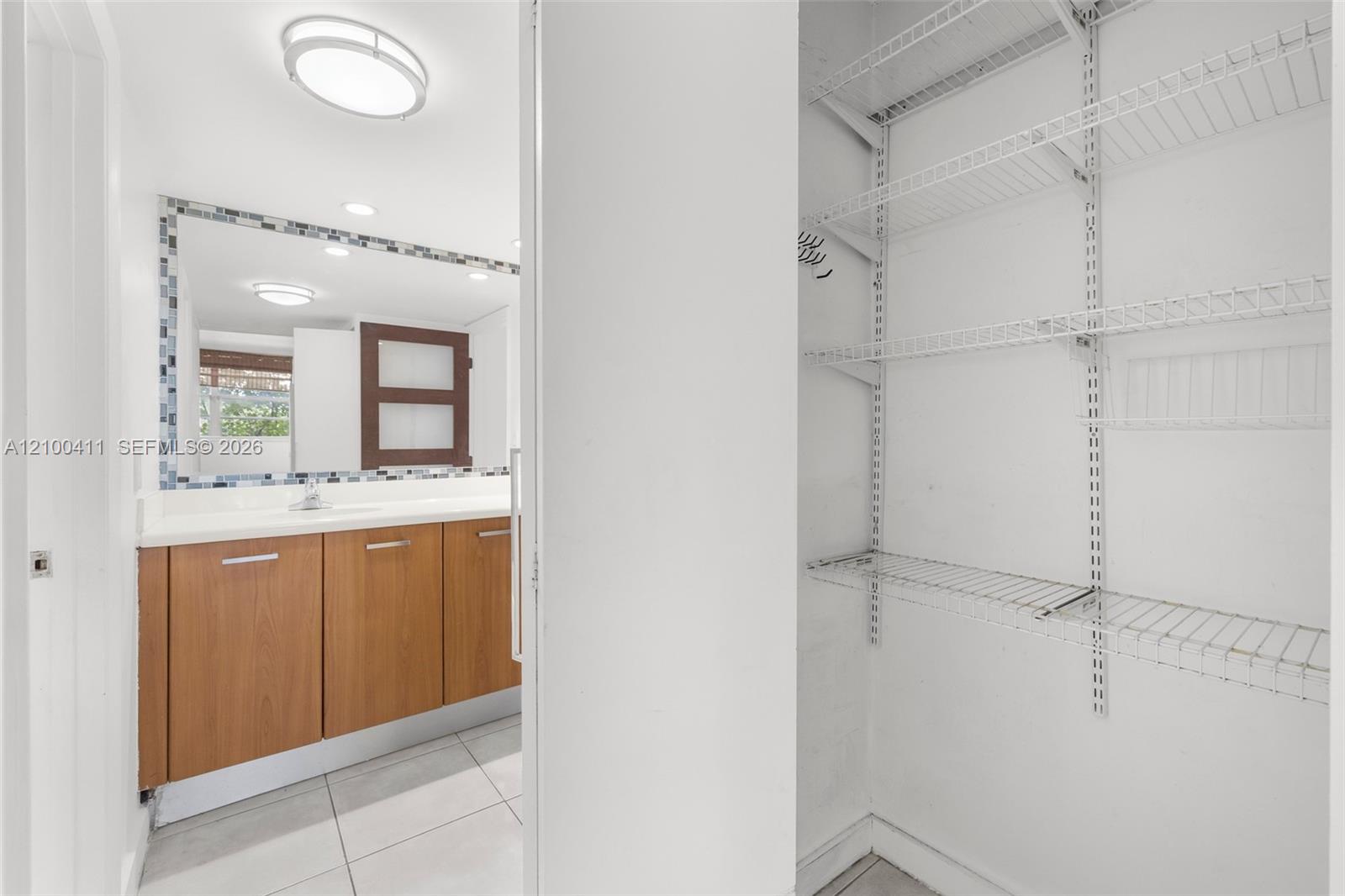 Bathroom featuring a wood-finish vanity with a solid-surface countertop, tiled flooring, a framed mirror with a decorative mosaic border, and a walk-in storage area with adjustable wire shelving