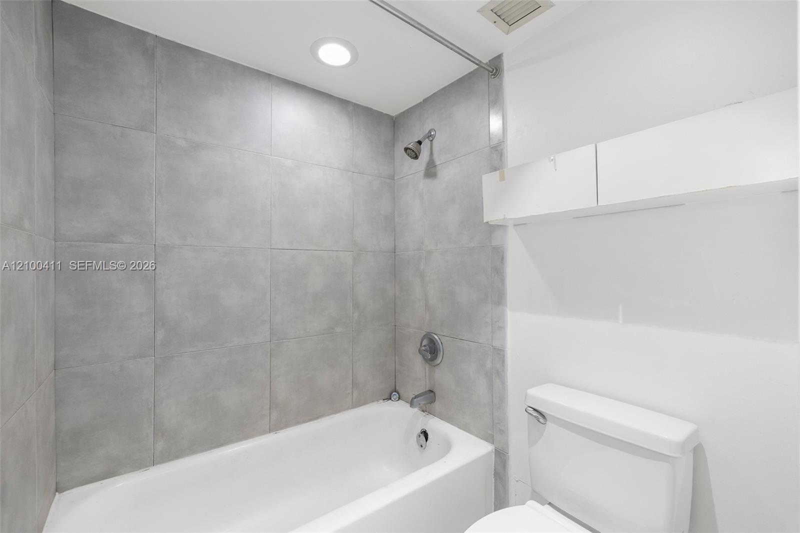 Full-height grey tile tub surround, white porcelain bathtub, wall-mounted shower head, recessed ceiling lighting, and built-in white upper cabinetry