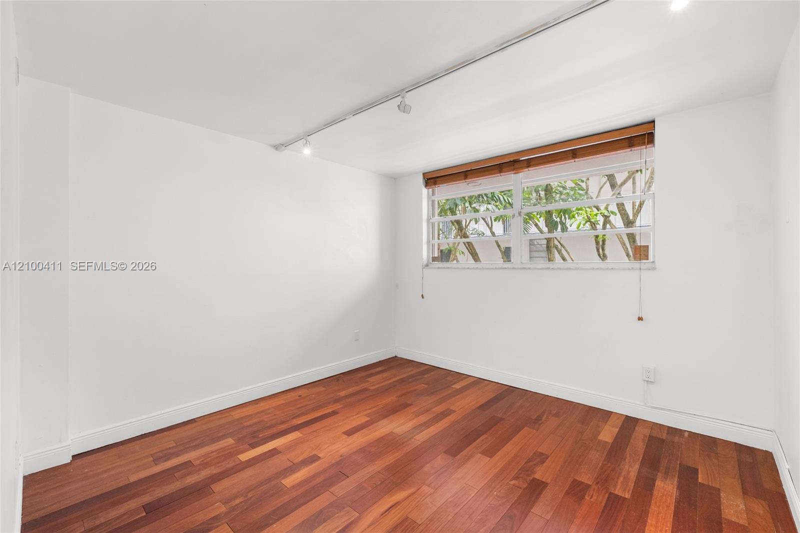 Interior room featuring wood-finish flooring, ceiling-mounted track lighting, and a horizontal window with slatted wood-tone blinds