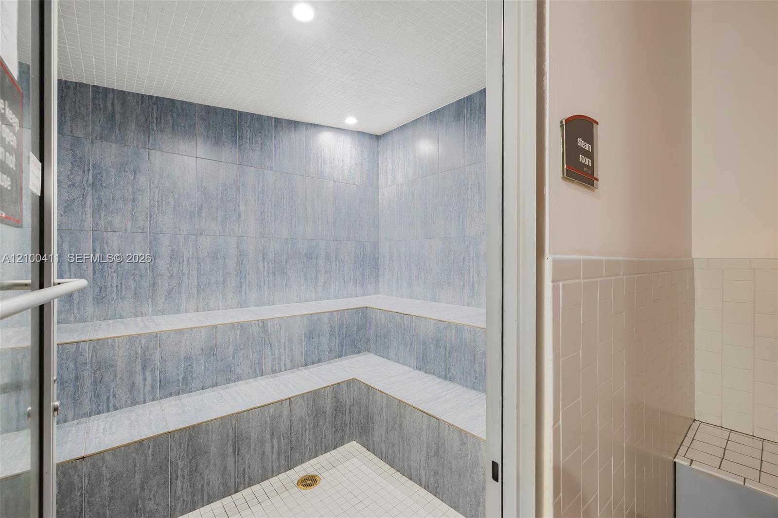 Steam room featuring full-height blue-toned tile wall cladding, tiered built-in bench seating, recessed lighting, and a white tiled floor