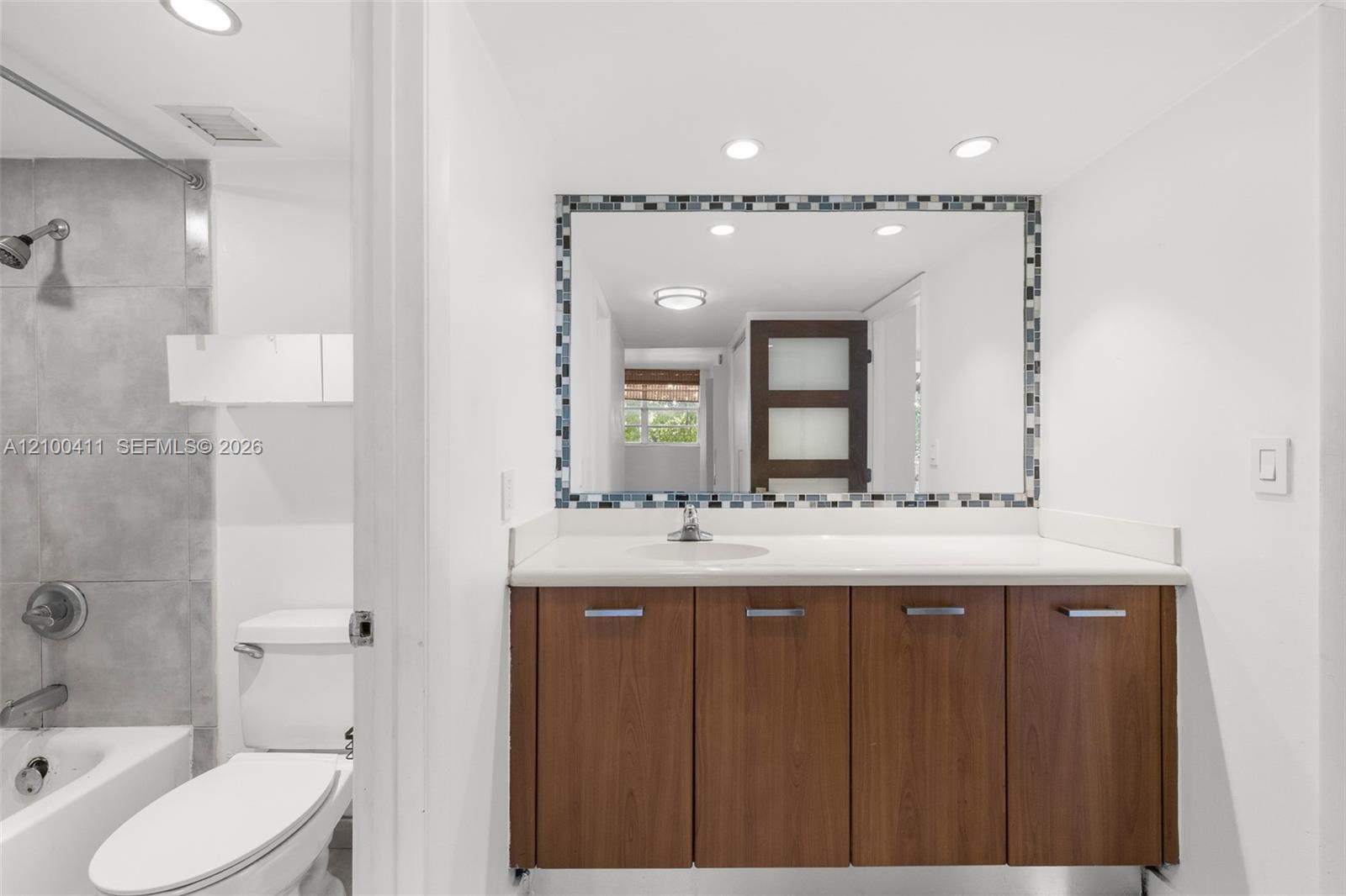 Vanity featuring wood-finish cabinetry, a white countertop with an integrated basin, a mosaic-tiled mirror frame, and recessed ceiling lighting