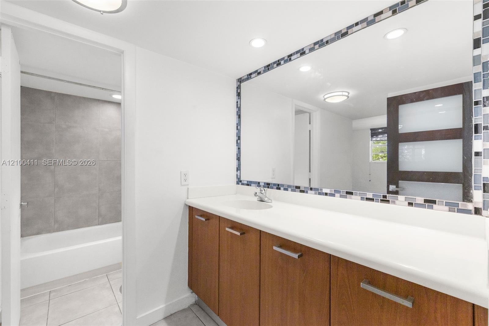 Bathroom featuring a wood-finish vanity with a solid-surface countertop, a large mirror with a decorative mosaic tile border, a tub-shower enclosure with oversized grey wall tile, and a multi-panel door with frosted glass inserts