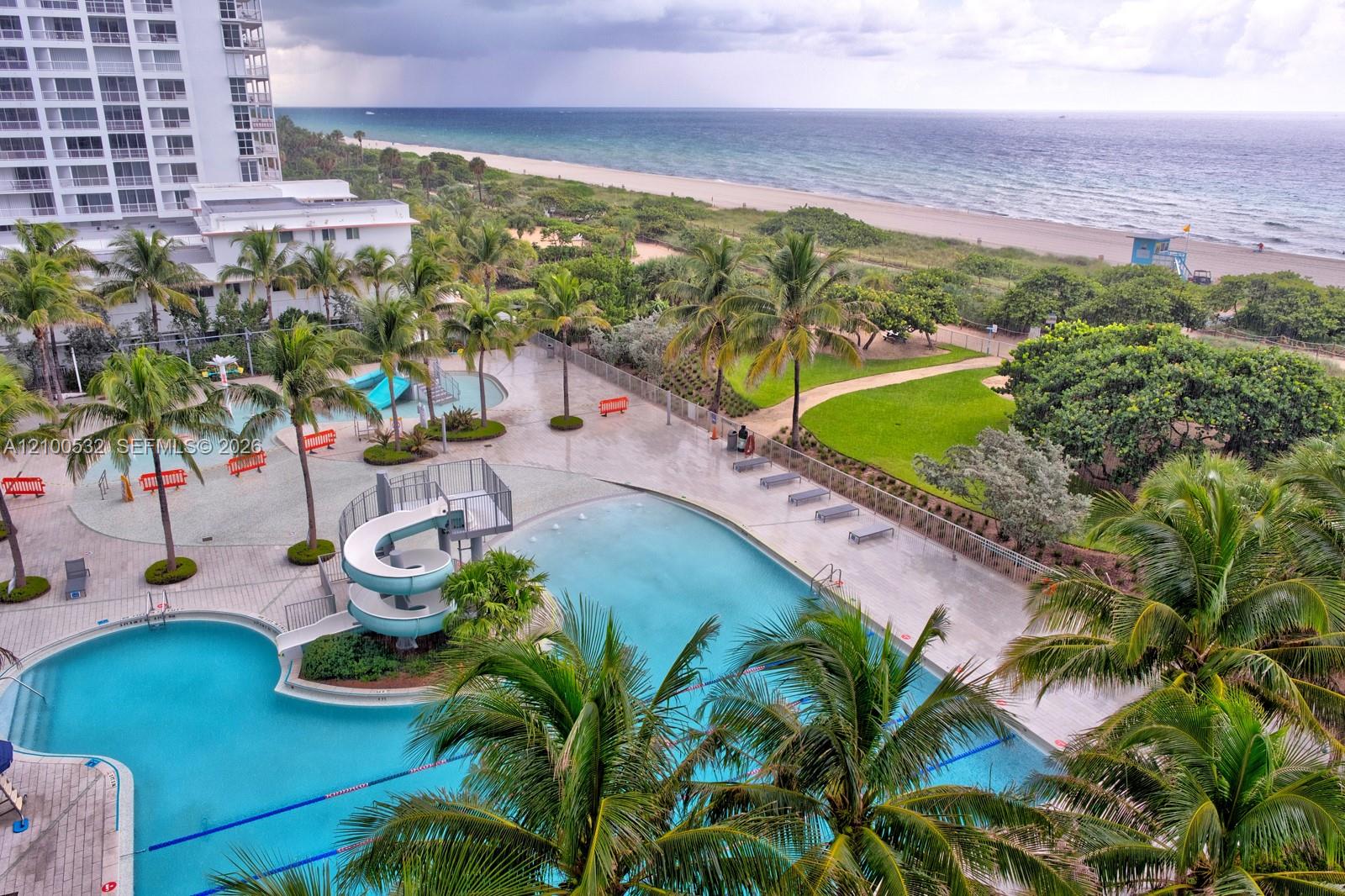 Outdoor swimming area featuring multiple pools, a water slide, expansive paved deck, and mature palm trees