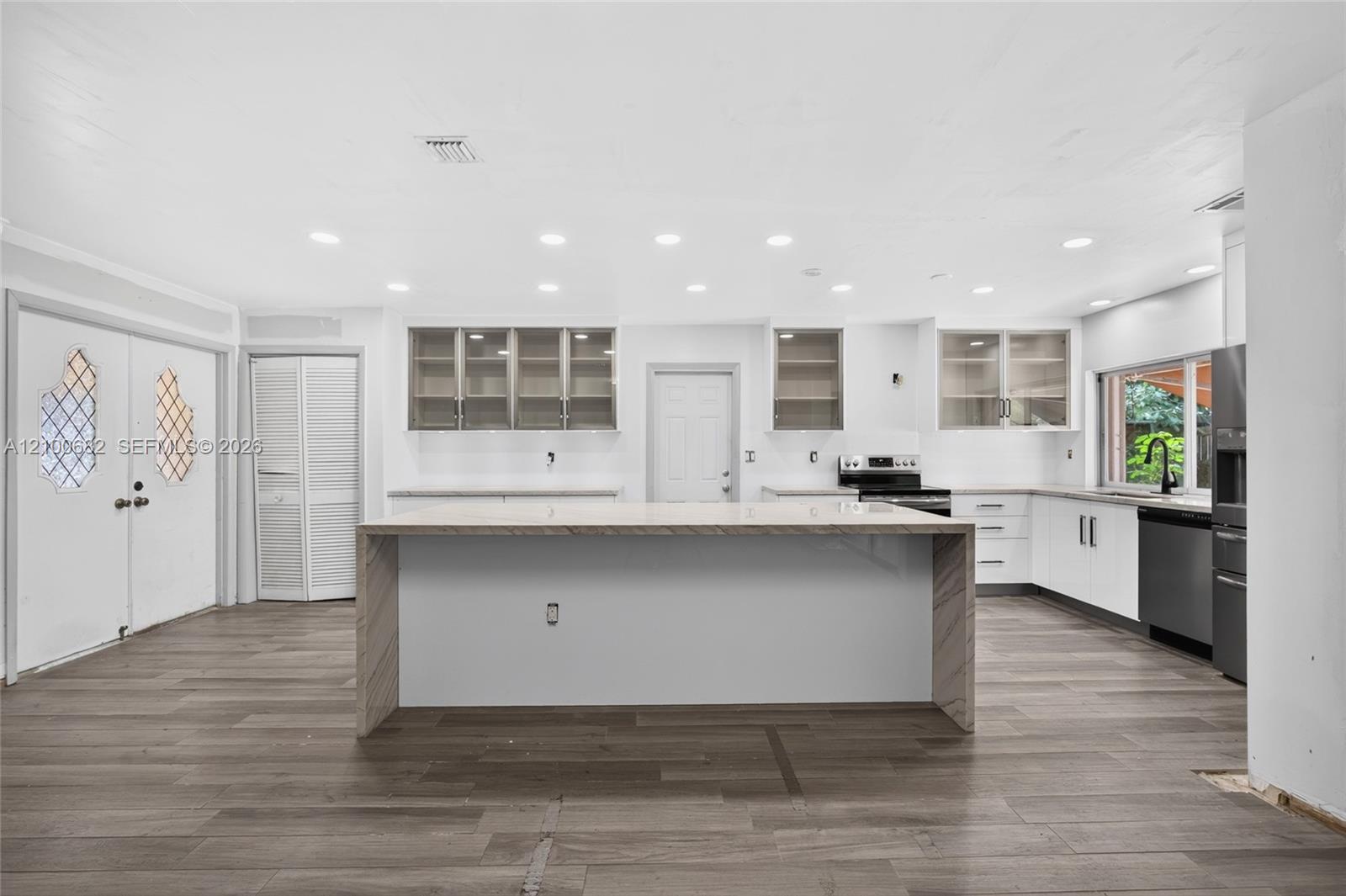 Kitchen featuring a waterfall-edge center island, vinly wood-finish flooring, white cabinetry with glass-front uppers, recessed lighting, and stainless steel appliances