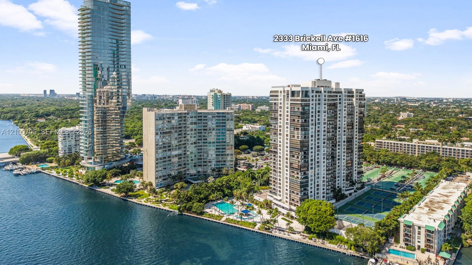 Aerial perspective of a multi-story residential high-rise featuring waterfront positioning, multiple outdoor swimming pools, and dedicated tennis courts