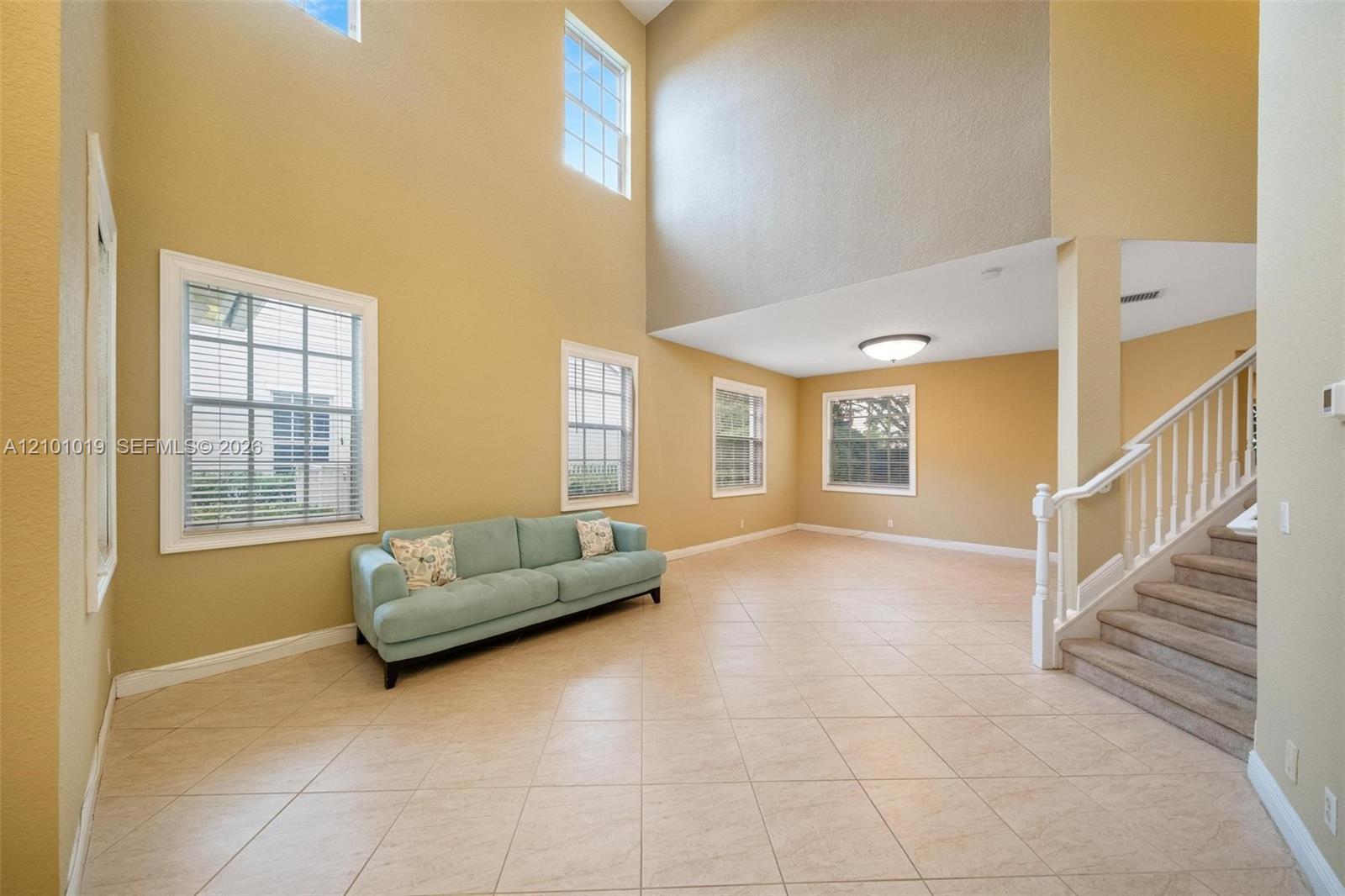 Expansive living area featuring vaulted ceilings, oversized windows for natural light, ceramic tile flooring, and a staircase with white balusters Expansive living area featuring vaulted ceilings, oversized windows for natural light, ceramic tile flooring, and a staircase with white balusters