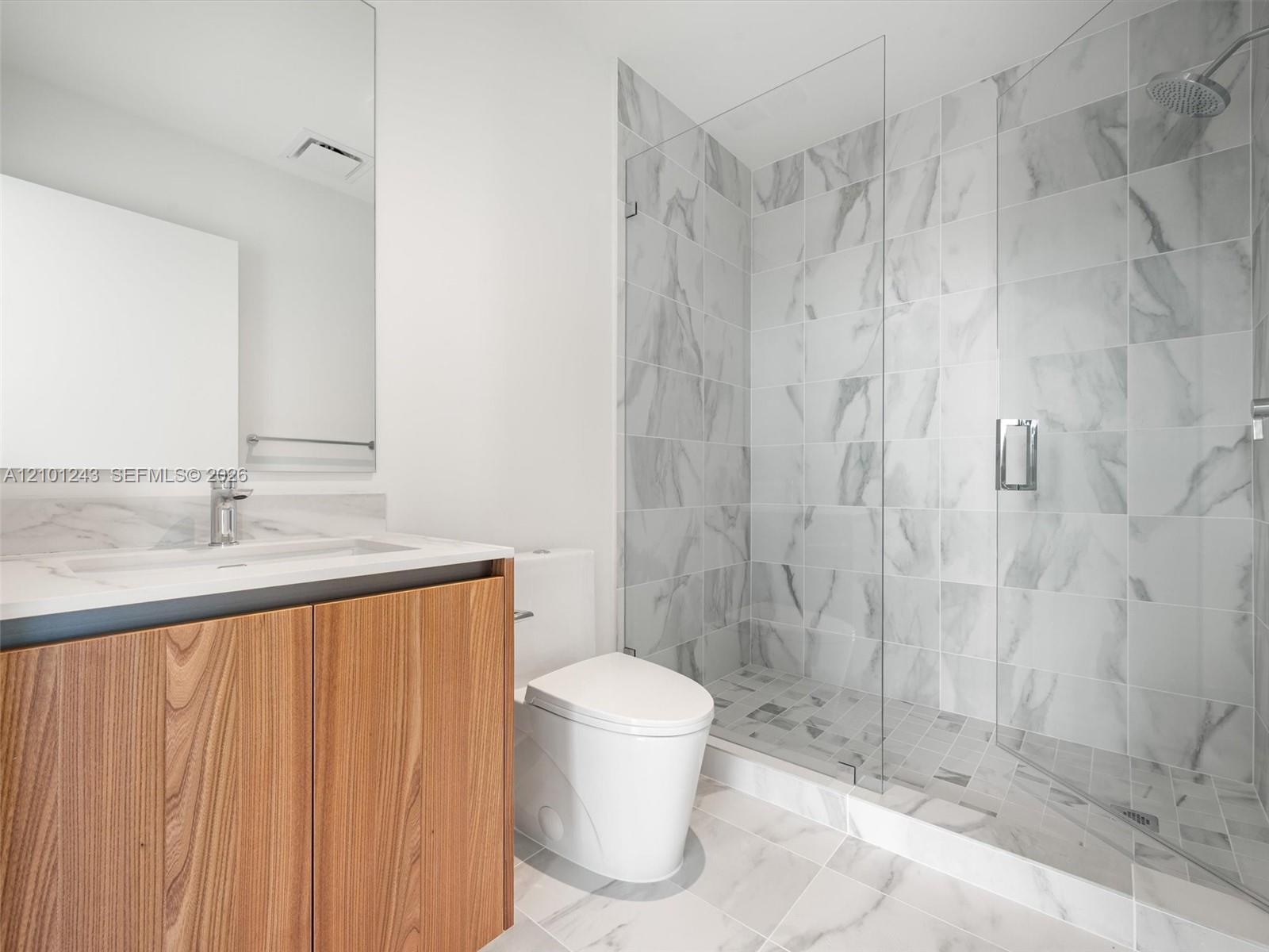 Bathroom featuring a wood-finish vanity with a stone-surfaced countertop, large-format tile flooring, walk-in glass-enclosed shower with floor-to-ceiling tile surround, and a wall-mounted mirror