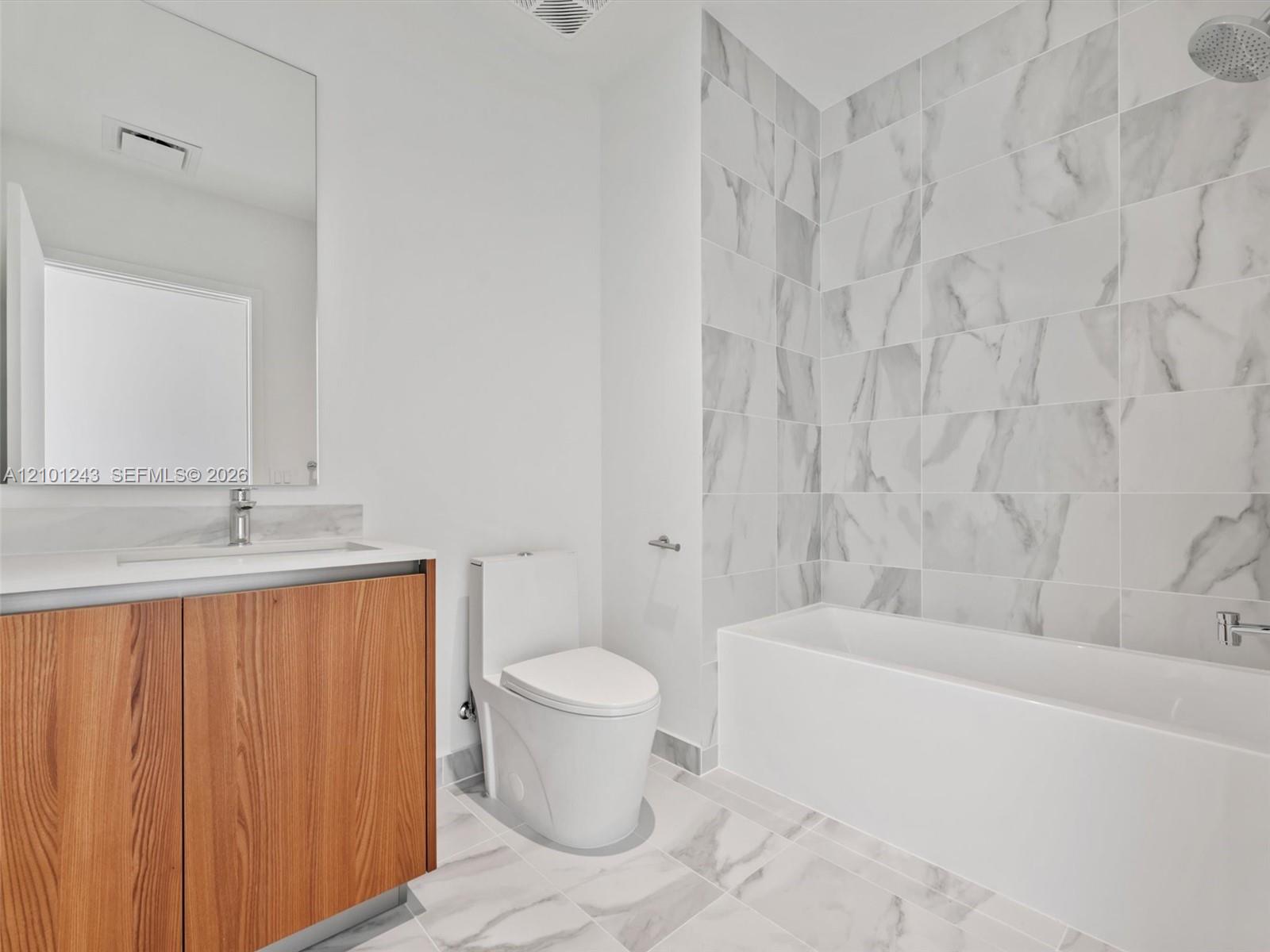 Bathroom featuring a wood-finish vanity with a stone-surface countertop, floor-to-ceiling tile shower surround, and large-format tile flooring