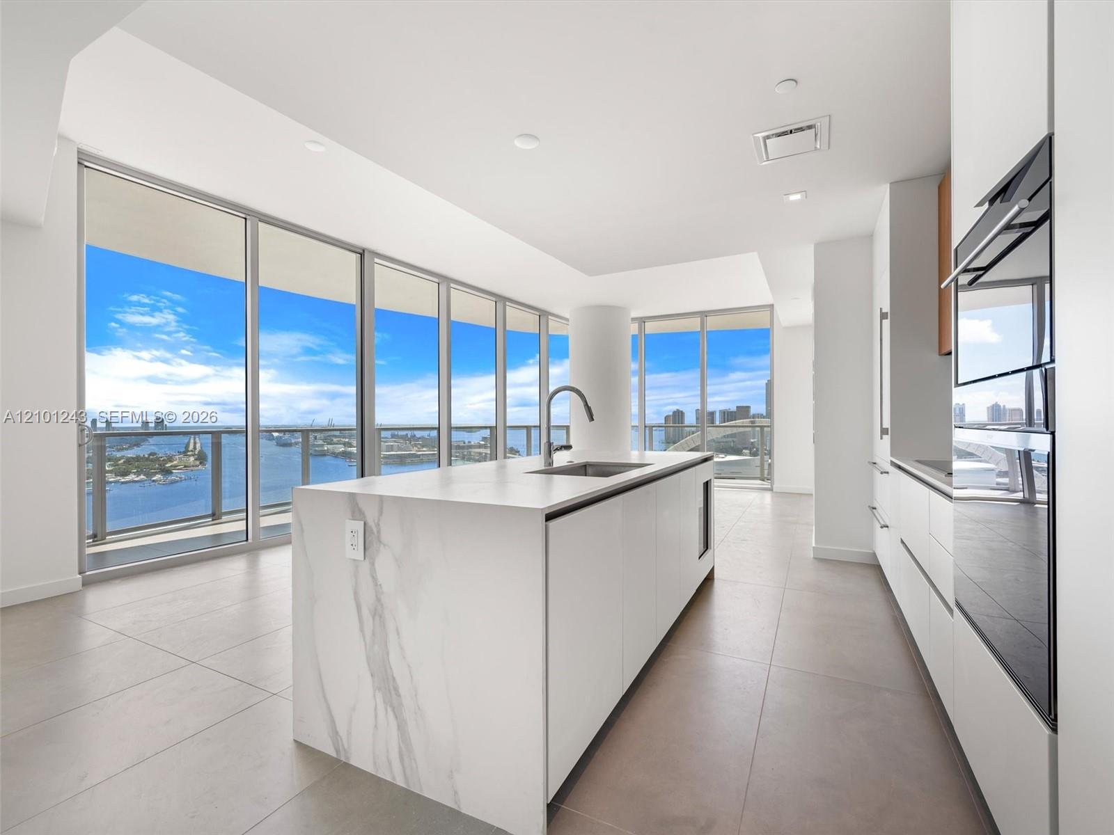 Kitchen featuring a central island with stone-finish waterfall edges, stainless steel under-mount sink, integrated appliances, floor-to-ceiling windows, and tile flooring