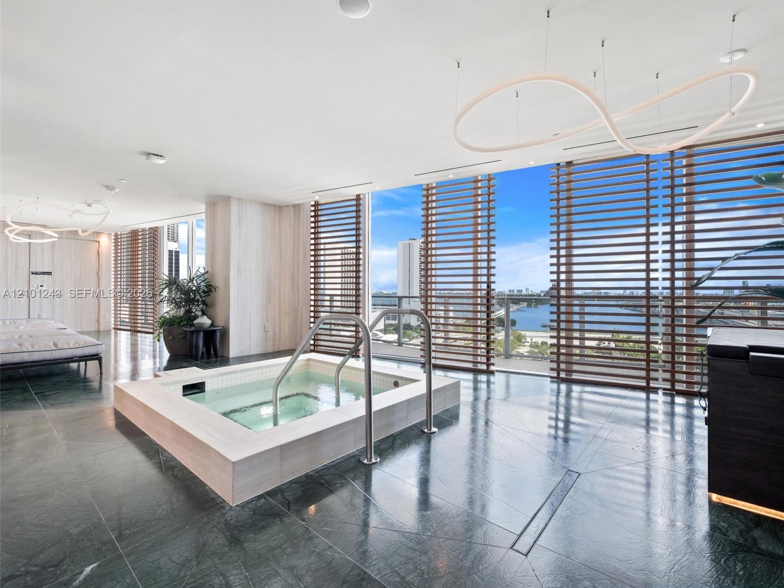 Built-in soaking tub with light-toned stone surround, polished dark stone flooring, floor-to-ceiling windows with horizontal wood-finish louvers, and architectural pendant lighting