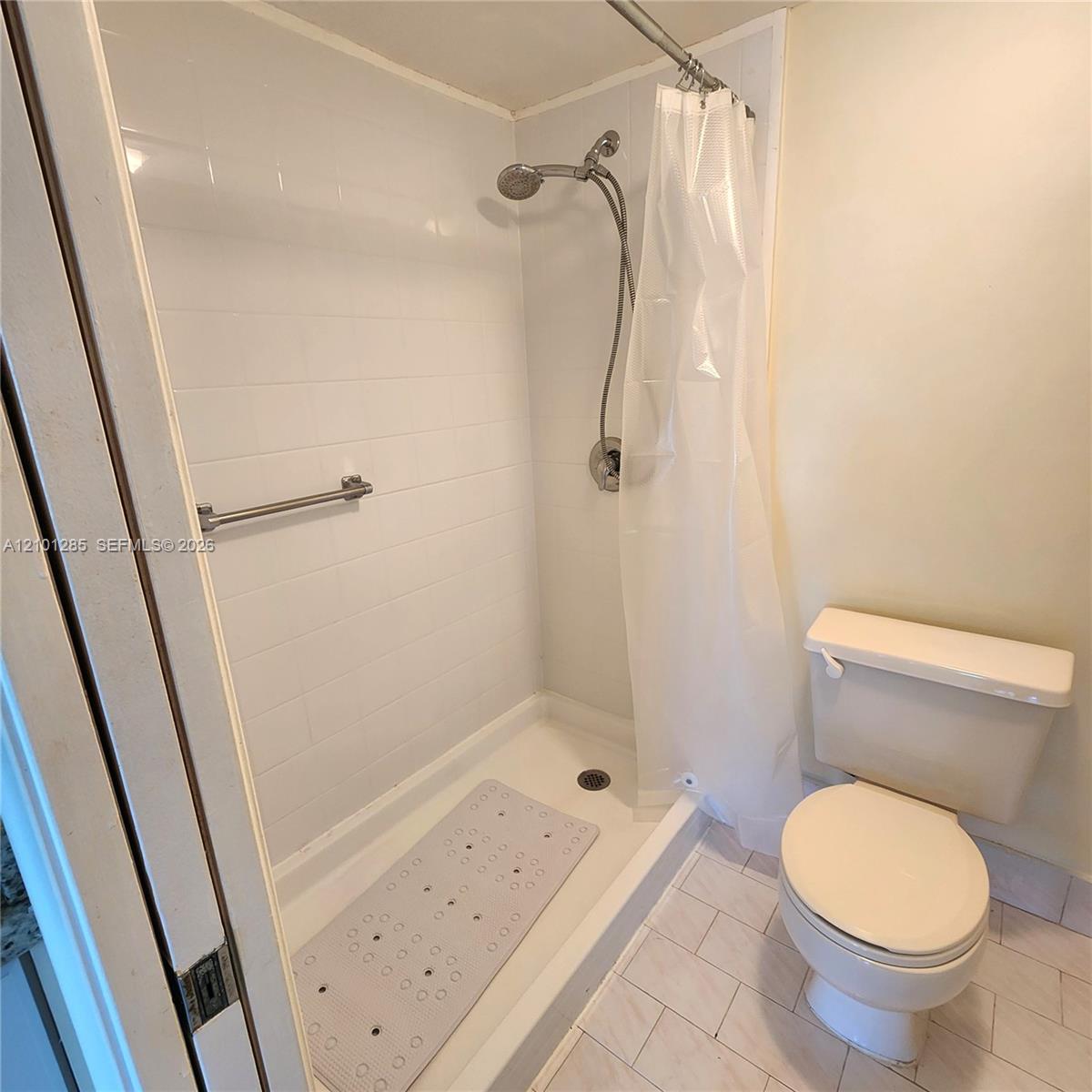 Bathroom featuring a walk-in shower with white tile surround, adjustable handheld showerhead, brushed metal grab bar, and neutral floor tile