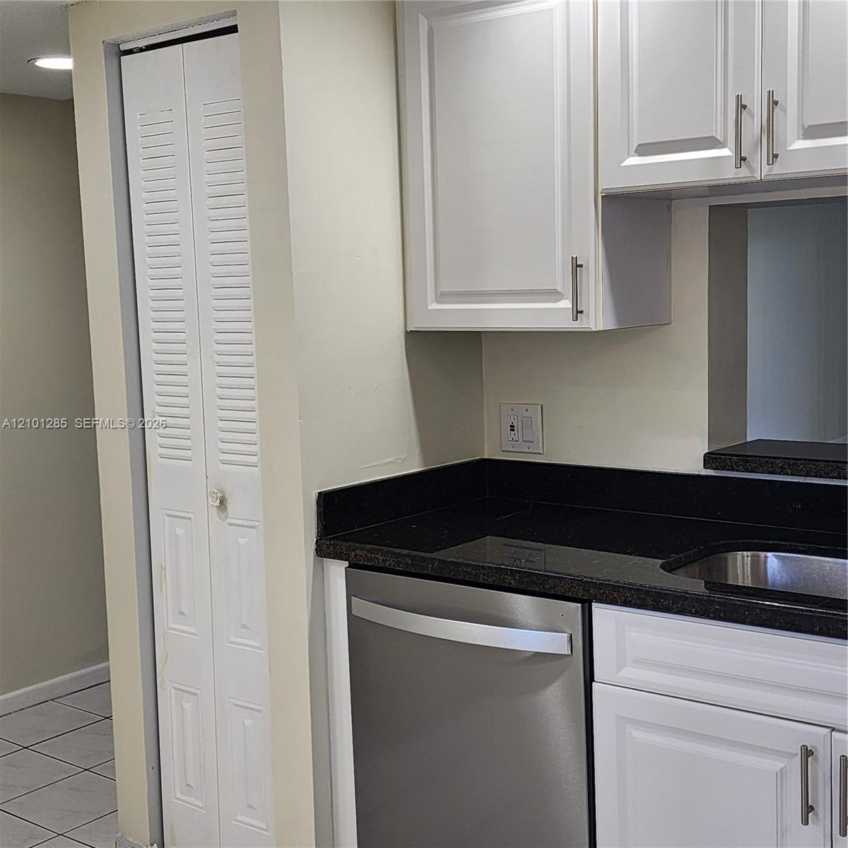 Kitchen featuring white cabinetry with brushed metal hardware, dark solid-surface countertops, a stainless steel dishwasher, and a full-height louvered door storage closet