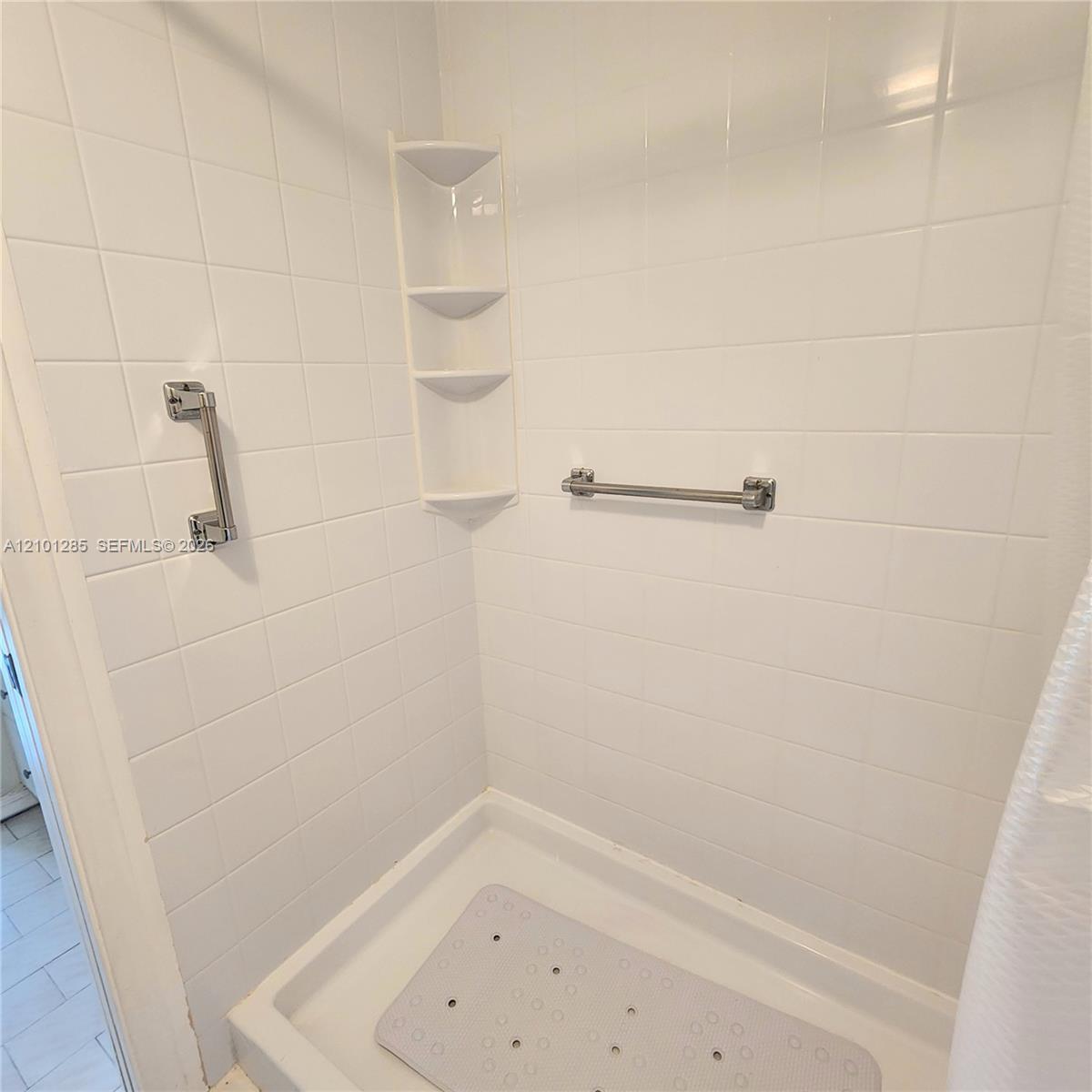 Shower stall featuring white square tile surround, built-in corner shelving, and dual stainless steel grab bars