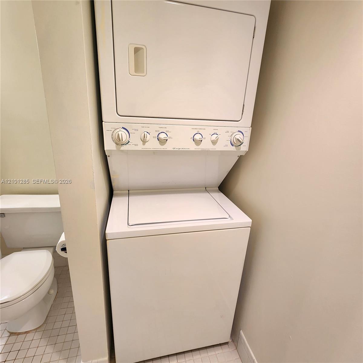 Stacked white laundry center featuring top-load washer and dryer configuration, paired with a white porcelain toilet on tiled flooring