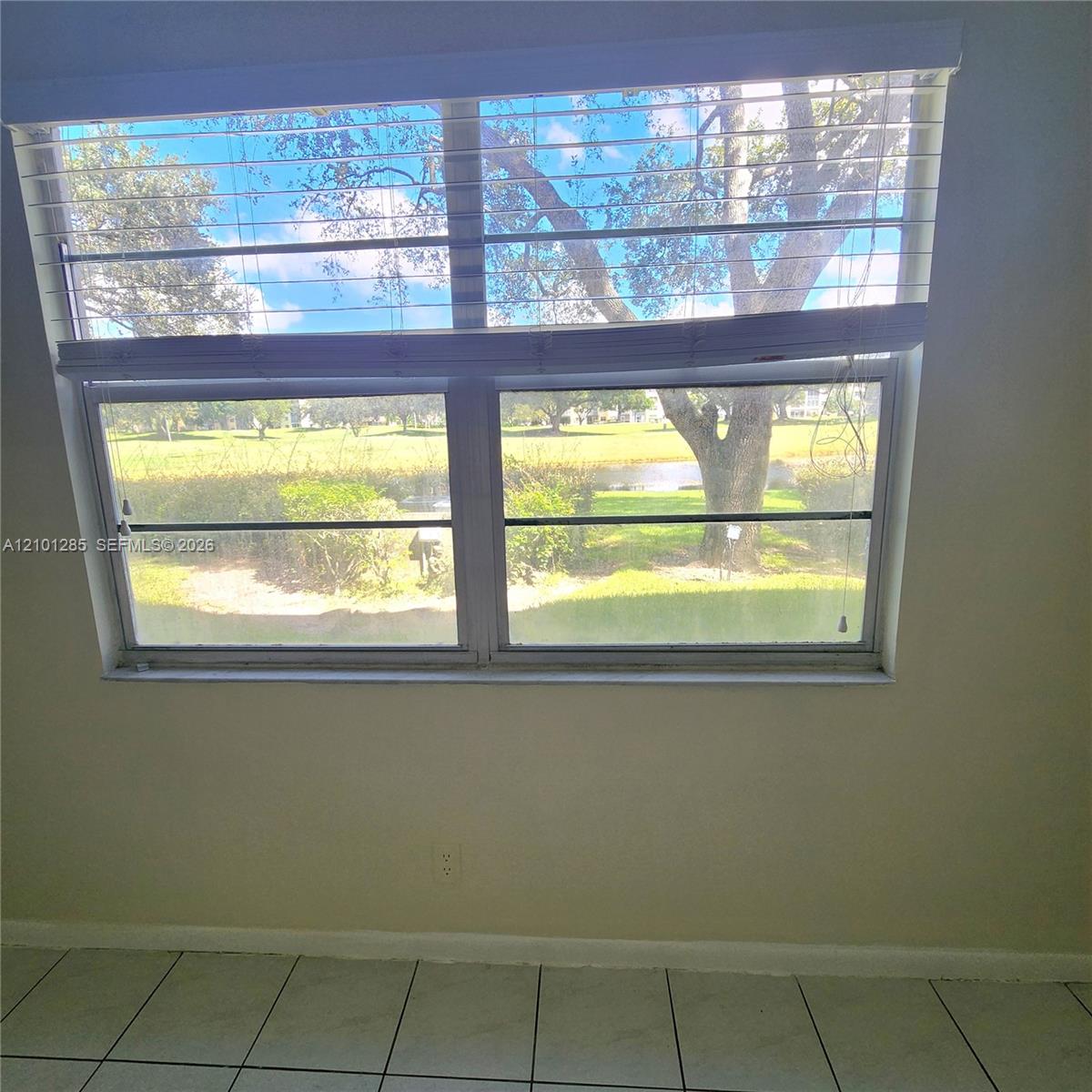 Large dual-pane window featuring a horizontal blind system, ceramic tile flooring, and exterior landscape scenery including mature trees and a pond
