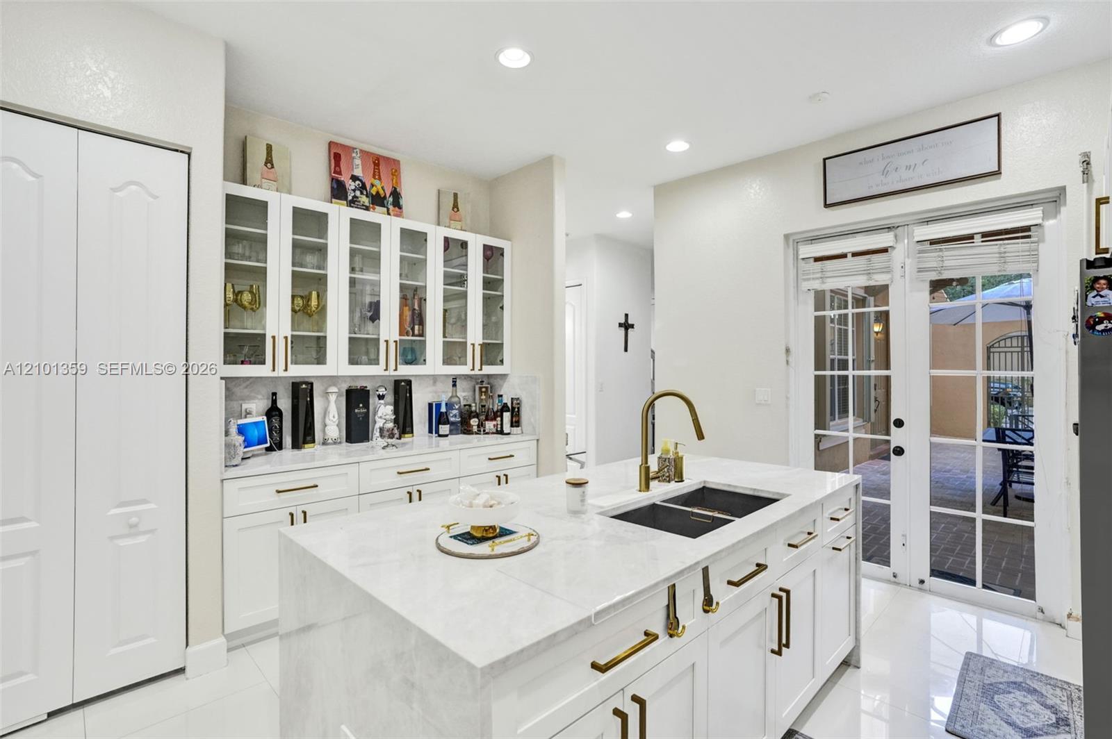 Kitchen island featuring a stone countertop, an undermount dual-basin sink, and a brass-finish faucet, complemented by built-in white cabinetry with glass-front display uppers, brass-finish hardware, and French doors