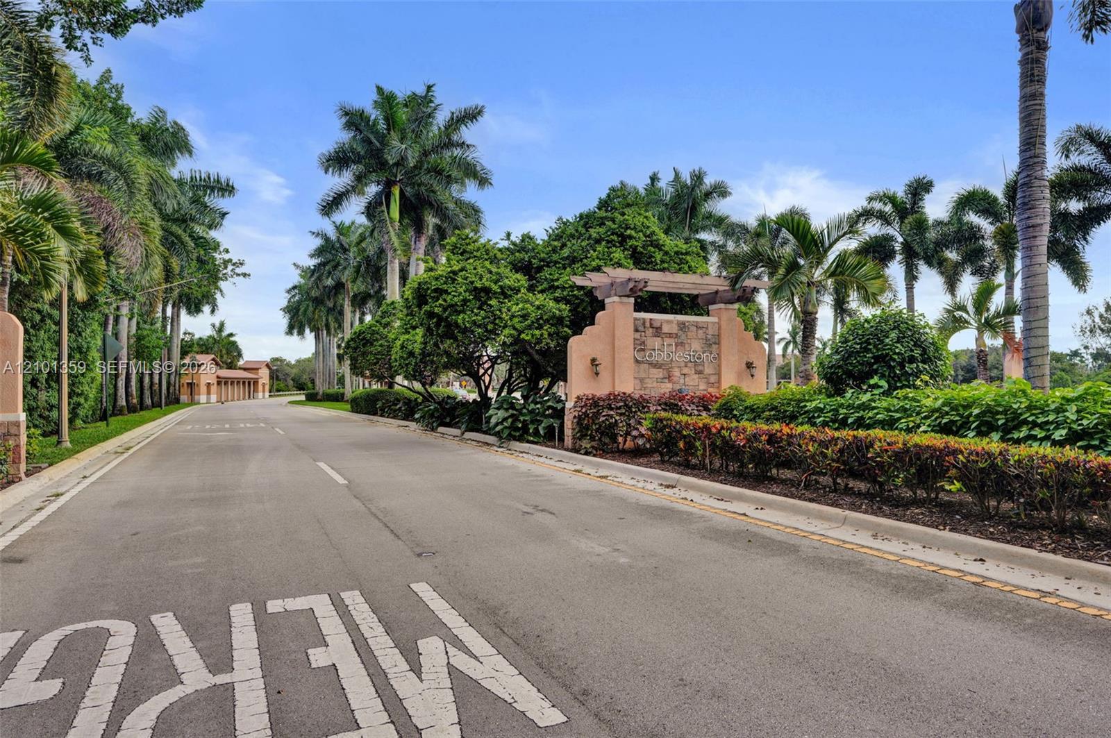 Stone-accented community monument signage featuring a wood-beam pergola, paved private roadway, and mature landscaping with established palm trees
