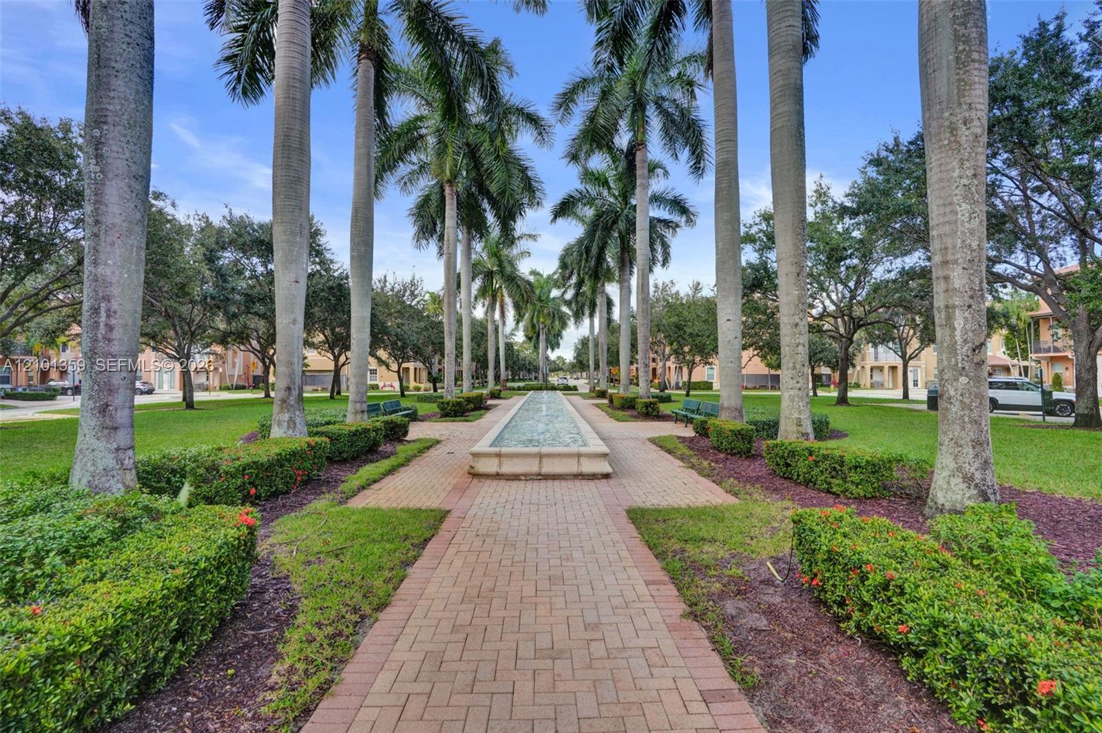Landscaped grounds featuring a central stone-clad fountain, brick-paved walkway, mature palm trees, manicured hedges, and expansive lawn areas