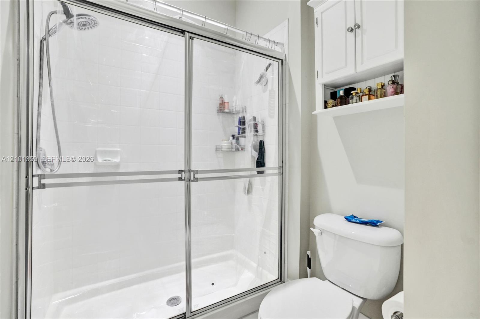 Walk-in shower with sliding glass doors, white subway-style tile surround, adjustable handheld shower head, and white built-in wall cabinetry
