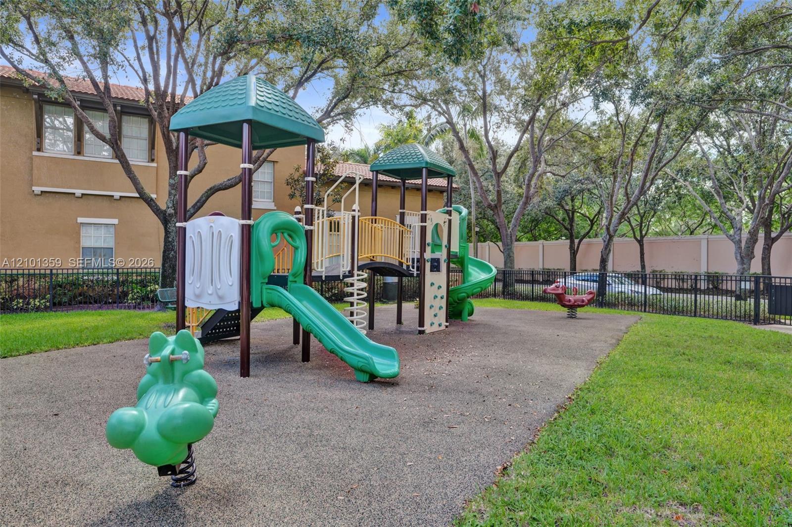 Recreational area featuring a multi-component play structure with slides and climbing elements, set within a landscaped perimeter enclosed by metal fencing and mature shade trees