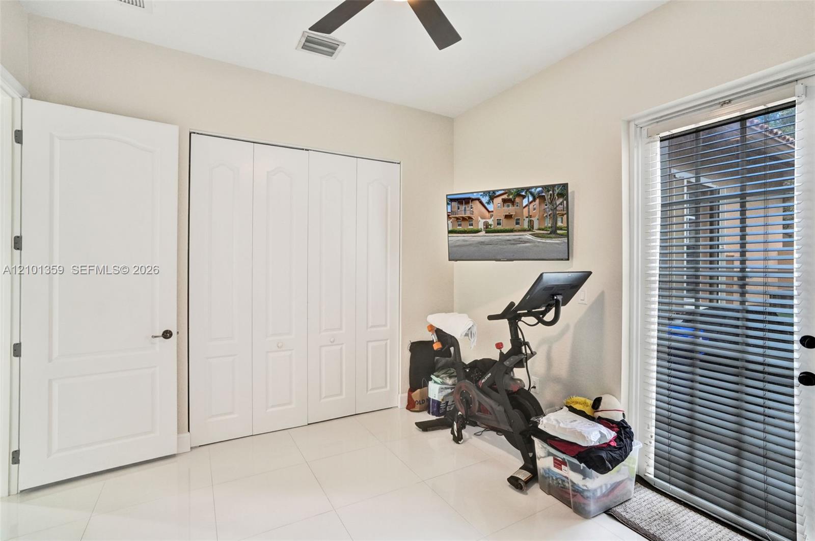 Neutral-toned room featuring large-format tile flooring, four-panel folding closet doors, a ceiling fan with integrated lighting, and a glass-paned door with horizontal blinds