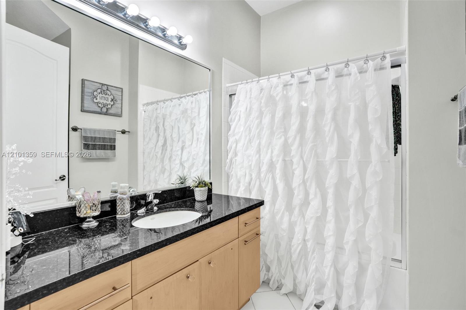 Full-width vanity with dark stone countertop, integrated oval basin, light-finish cabinetry, oversized wall-to-wall mirror, and multi-bulb vanity lighting