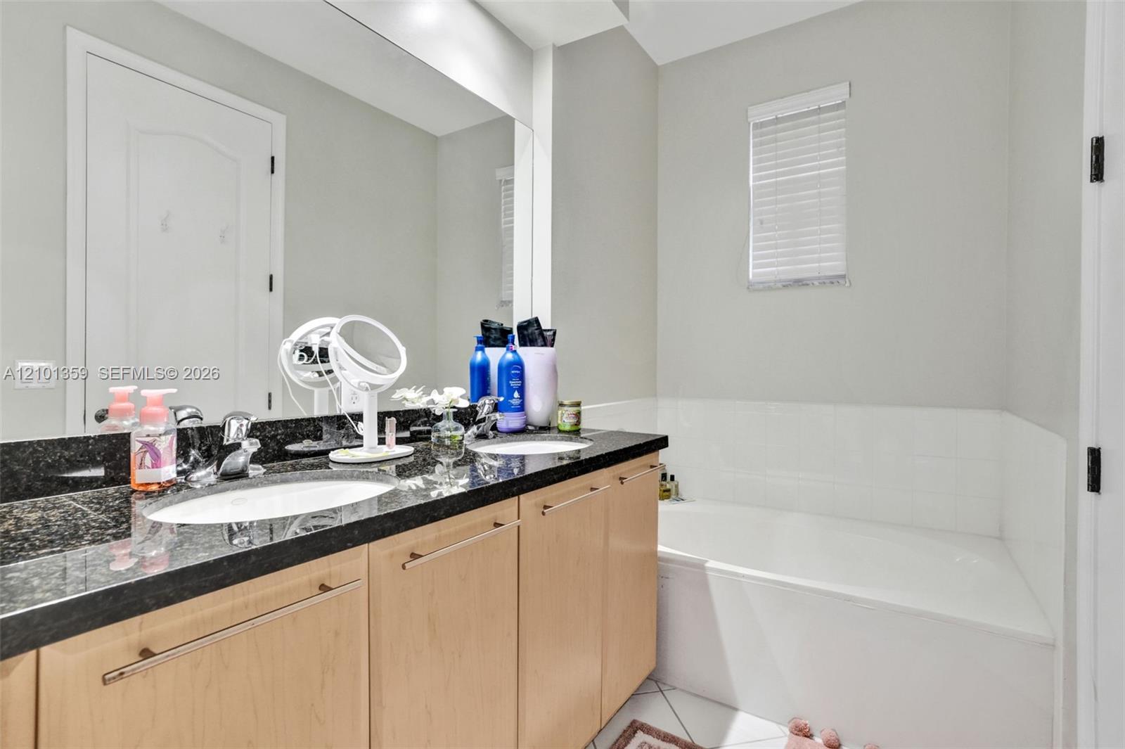 Dual-sink vanity with dark stone countertop, light-wood cabinetry, large wall-mounted mirror, and tile-surround bathtub
