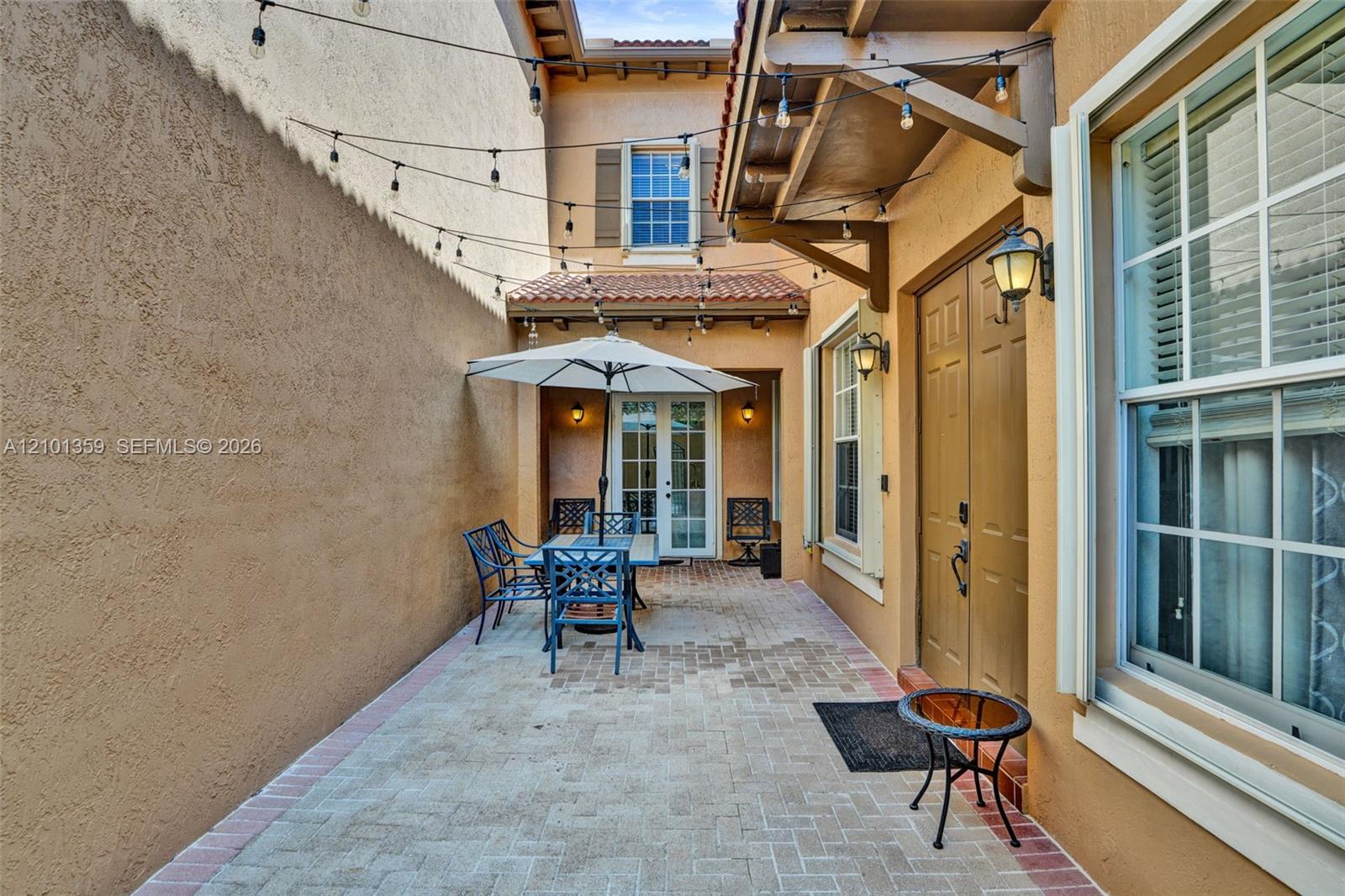 Enclosed courtyard featuring herringbone-patterned paver flooring, stucco wall finish, terracotta-style roof accents, and double-door entry with exterior sconce lighting