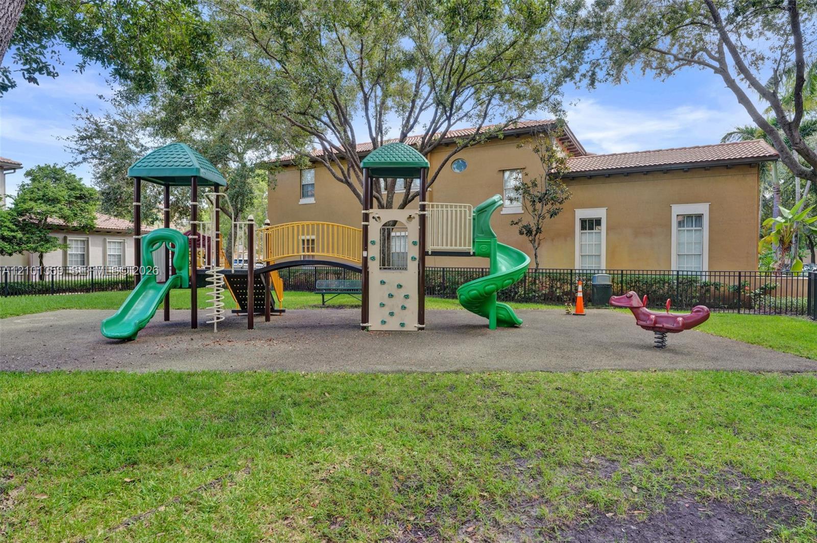 Community playground featuring a multi-level structure with two slides, a bridge, climbing elements, and a spring rider set within a grass-covered area enclosed by black metal fencing