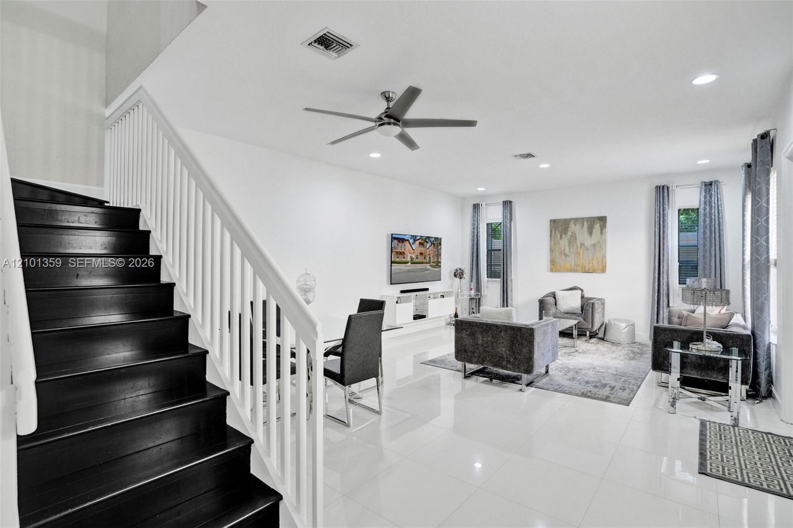 Open-concept interior featuring large-format tile flooring, a staircase with dark-finished treads and white handrails, recessed ceiling lighting, and a modern ceiling fan