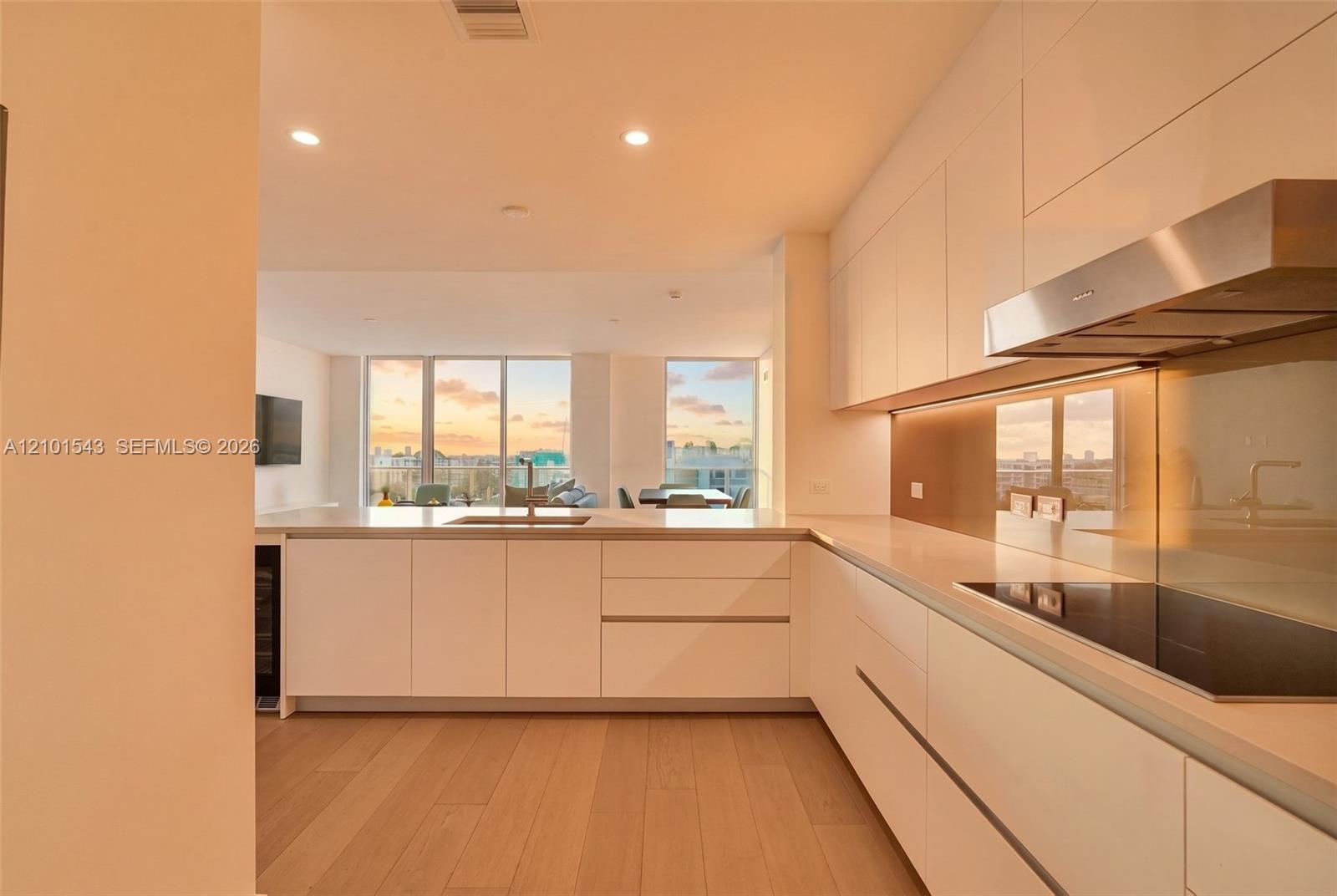 Contemporary kitchen featuring flat-panel cabinetry, light-toned wood-finish flooring, a stainless steel range hood, an integrated cooktop, and floor-to-ceiling windows Contemporary kitchen featuring flat-panel cabinetry, light-toned wood-finish flooring, a stainless steel range hood, an integrated cooktop, and floor-to-ceiling windows