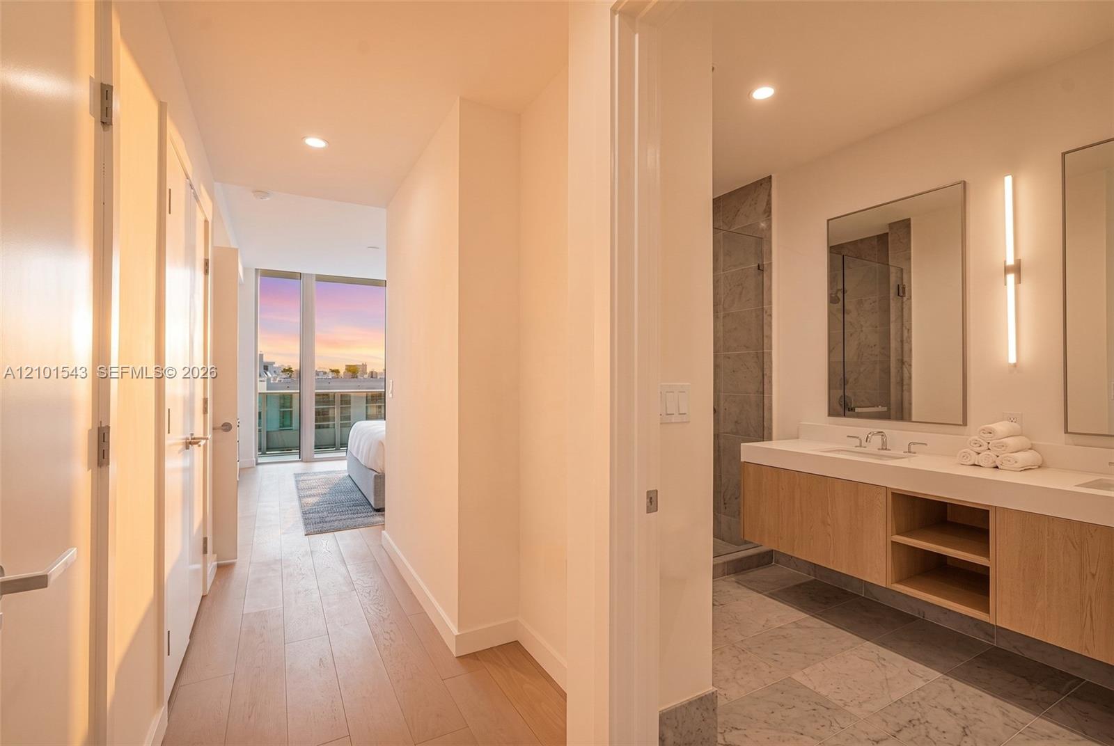 Floating wood-finish vanity with dual sinks, stone flooring, walk-in tile shower with glass enclosure, recessed lighting, and wall-mounted vanity mirrors