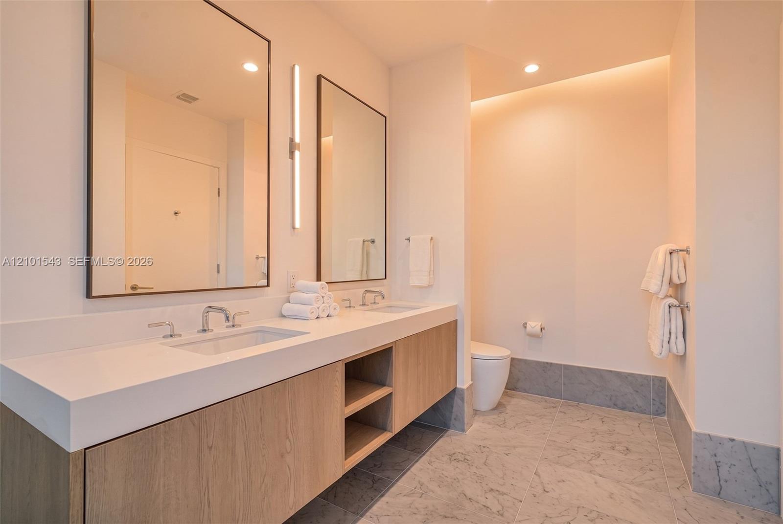 Dual-sink floating vanity with light wood-finish cabinetry, crisp stone countertop, integrated vertical wall lighting, and natural stone flooring