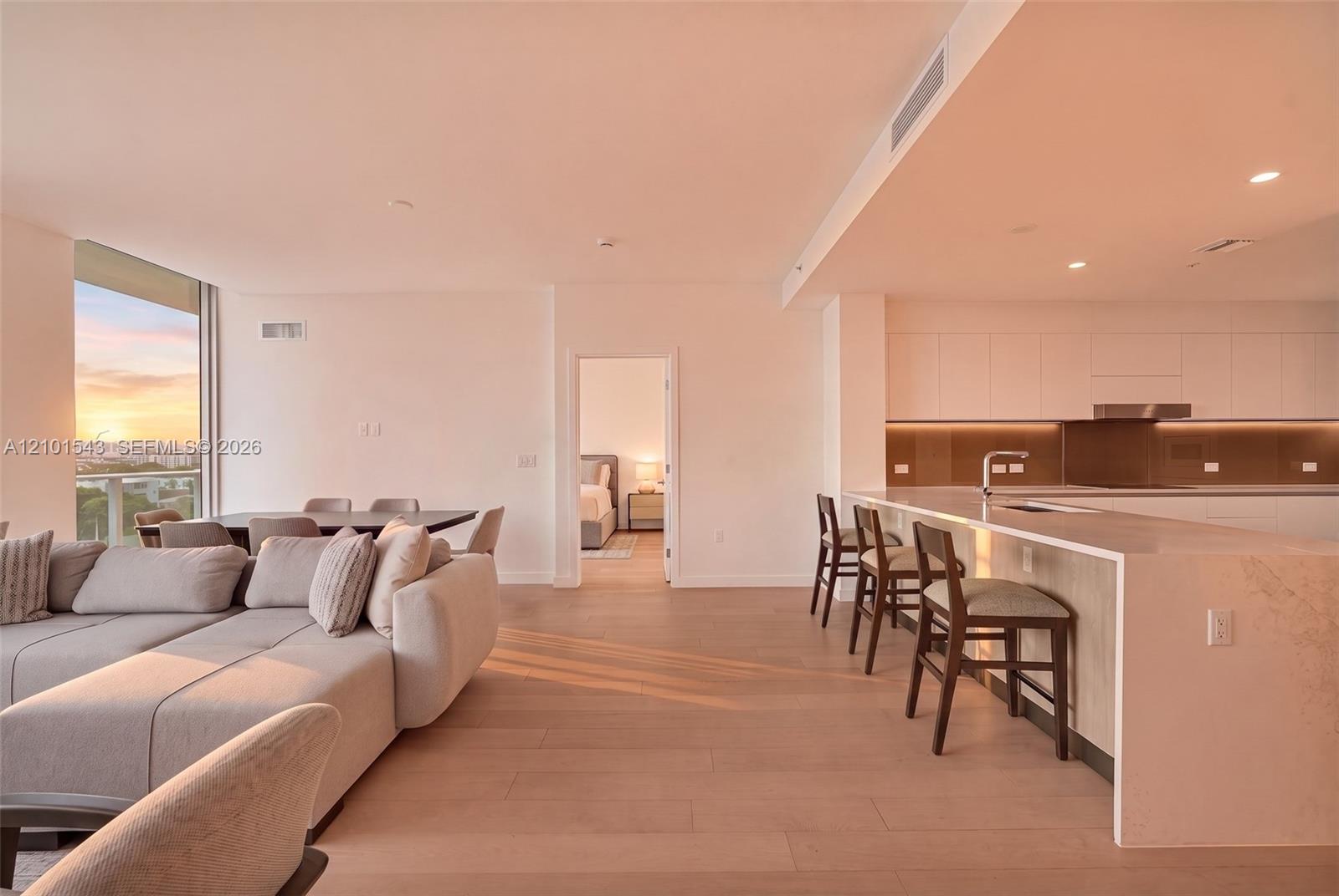 Open-concept interior featuring light wood-finish flooring, a kitchen with flat-panel cabinetry, a waterfall-edge countertop island, and recessed ceiling lighting Open-concept interior featuring light wood-finish flooring, a kitchen with flat-panel cabinetry, a waterfall-edge countertop island, and recessed ceiling lighting