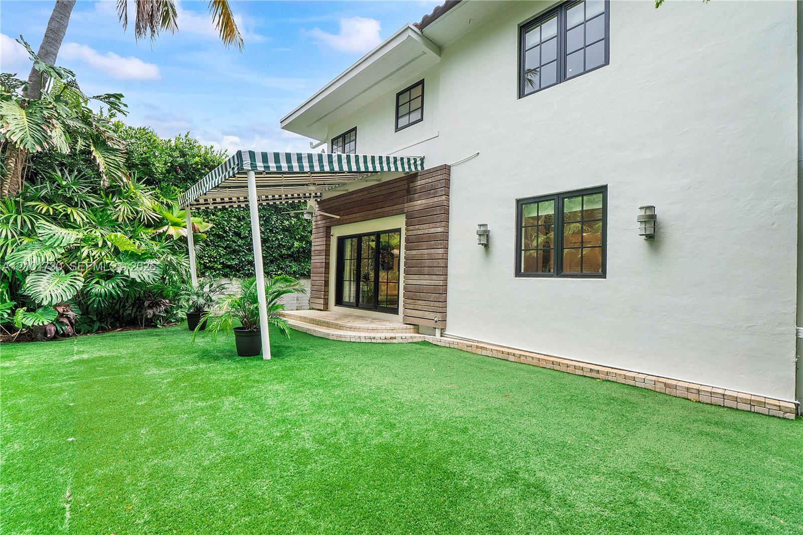 Exterior space featuring a manicured lawn, tiered stone patio, attached striped awning, wood-slat siding accents, and multi-pane windows