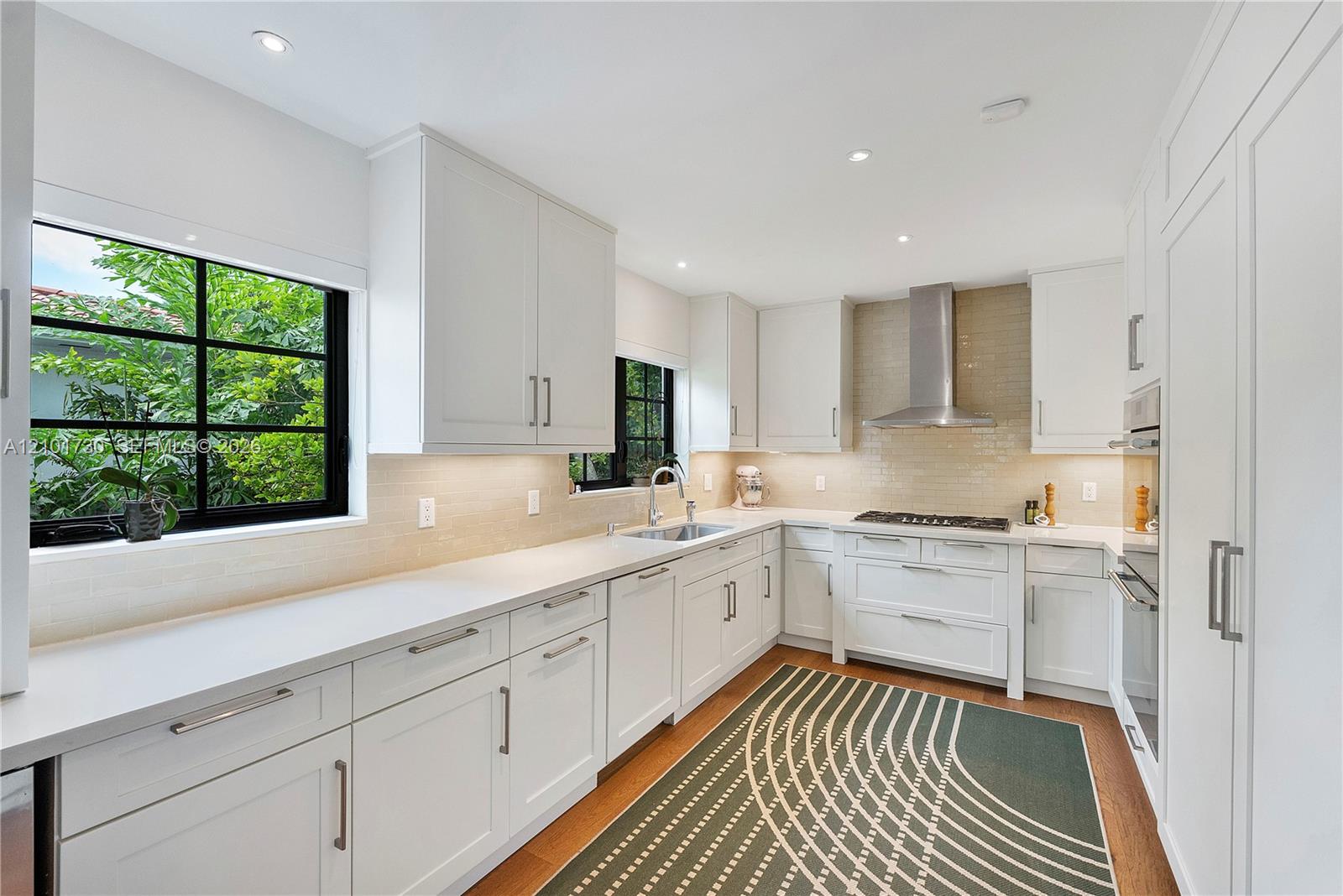 Kitchen featuring white shaker-style cabinetry, quartz countertops, subway tile backsplash, stainless steel ventilation hood, and wood-finish flooring