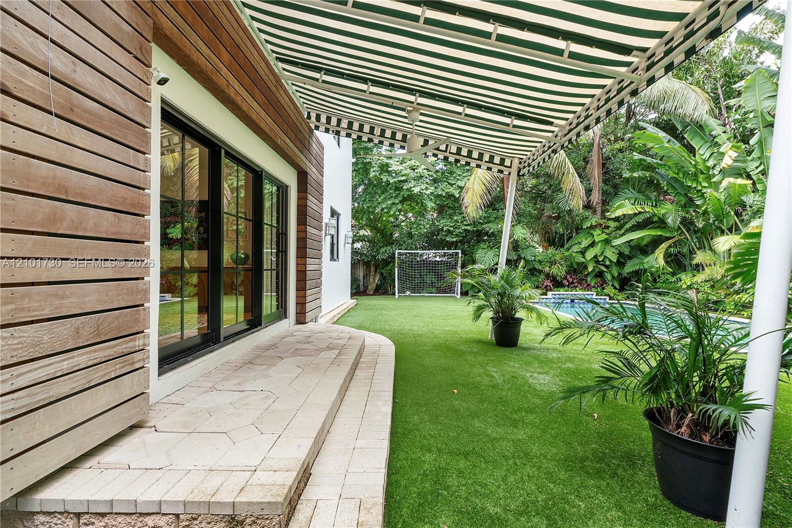 Exterior features include horizontal wood-slat siding, a retractable striped awning, a stone-paver patio, a swimming pool, and artificial turf lawn