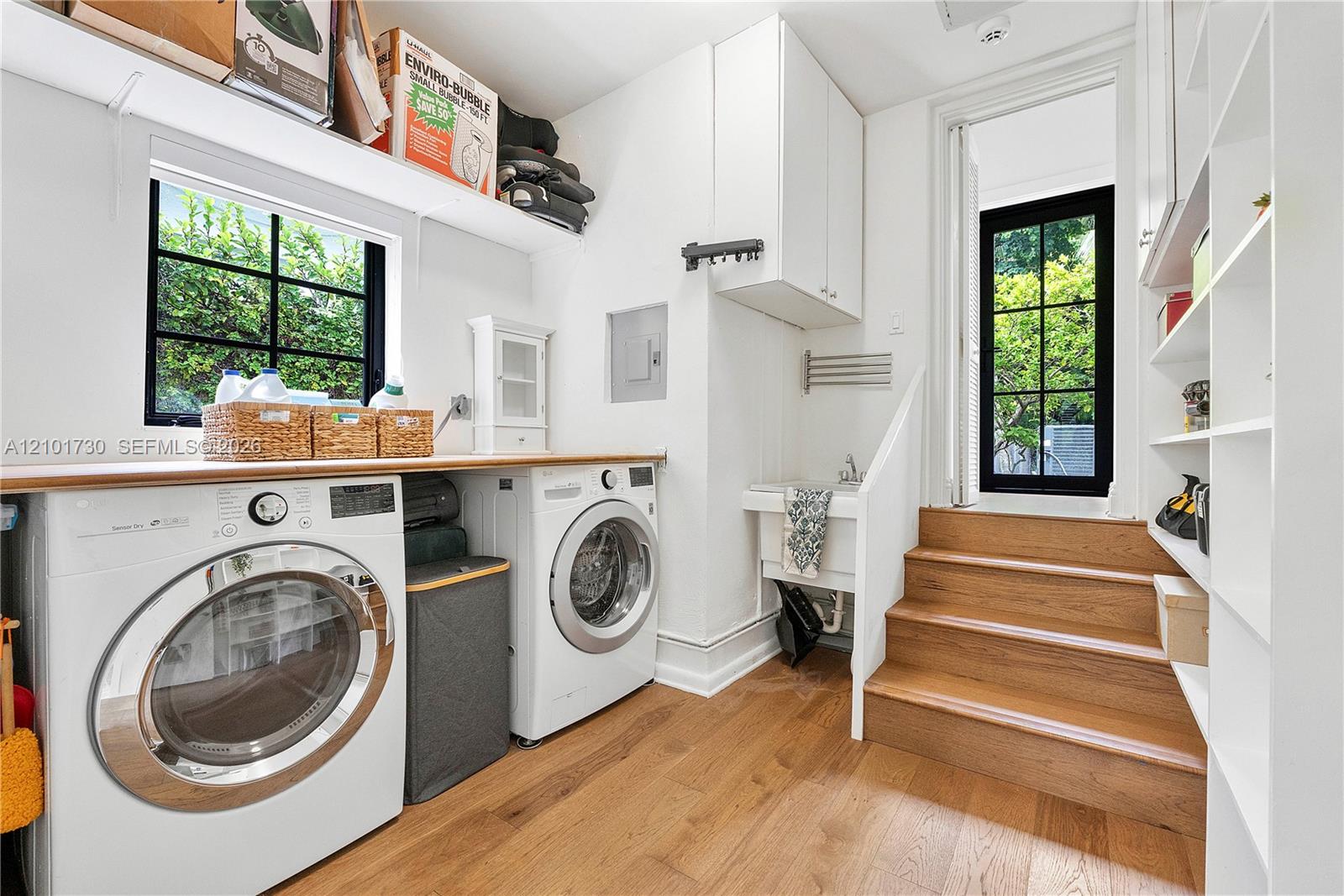 Laundry area featuring wood-finish flooring, built-in shelving, white cabinetry, a utility sink, and black-framed grid windows