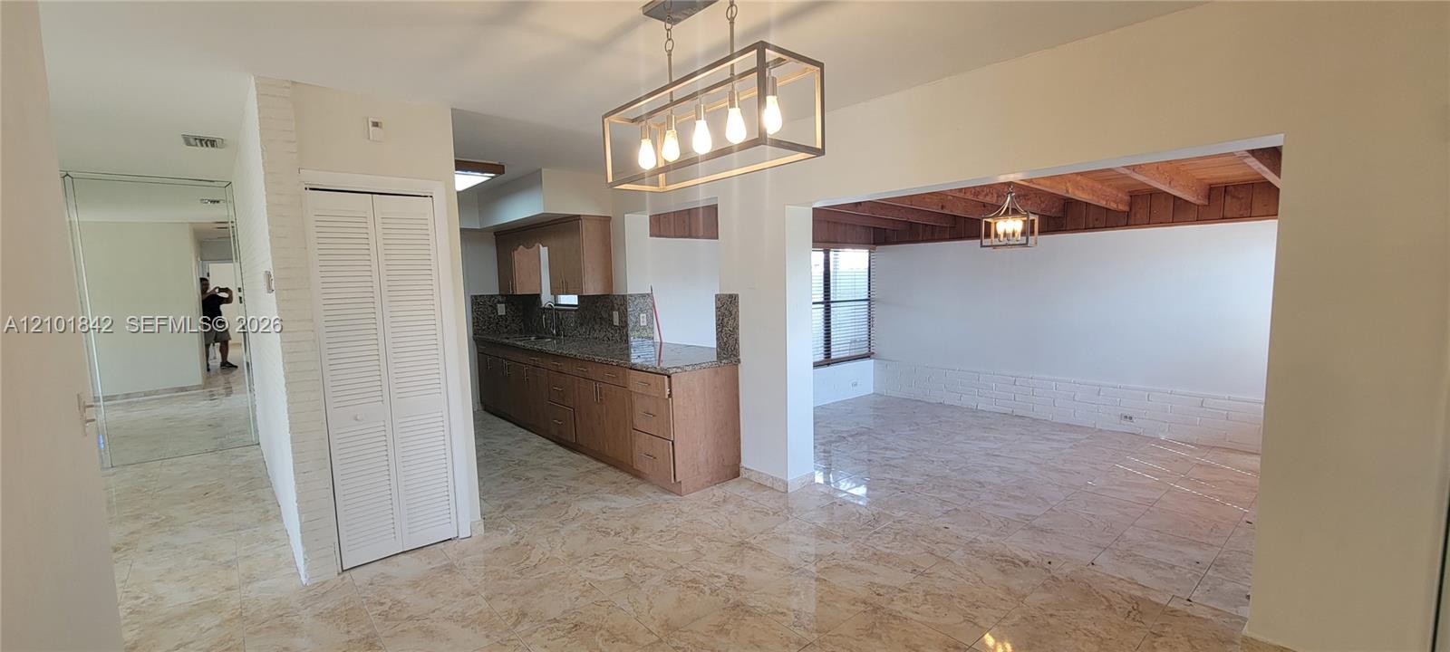 Open-concept interior featuring large-format tile flooring, a kitchen with stone countertops and wood cabinetry, exposed wooden ceiling beams, and a rectangular metal-frame pendant light fixture