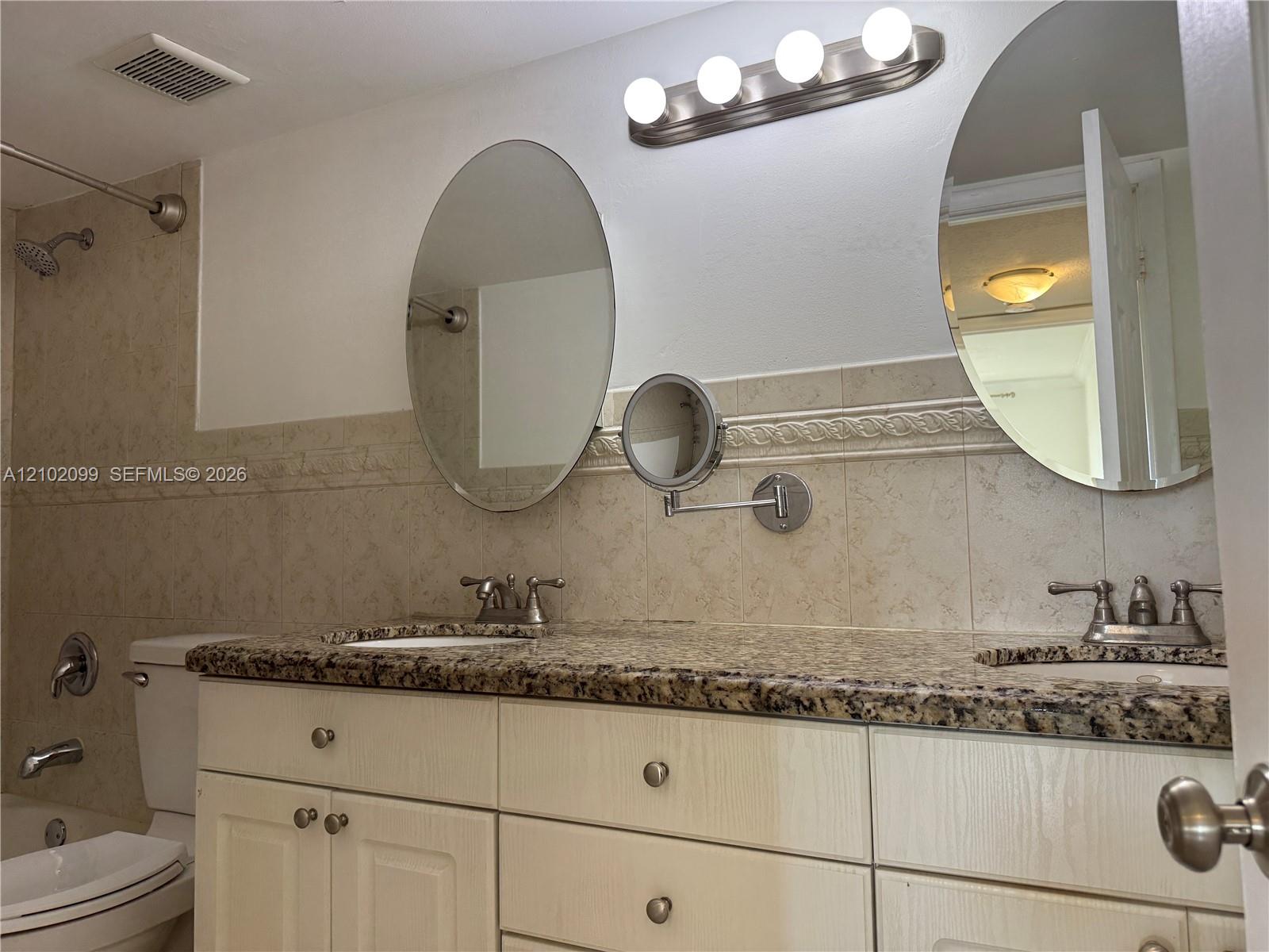 Double vanity featuring a stone countertop with dual undermount sinks, light-finish cabinetry, two oval wall-mounted mirrors, and an adjustable magnifying vanity mirror