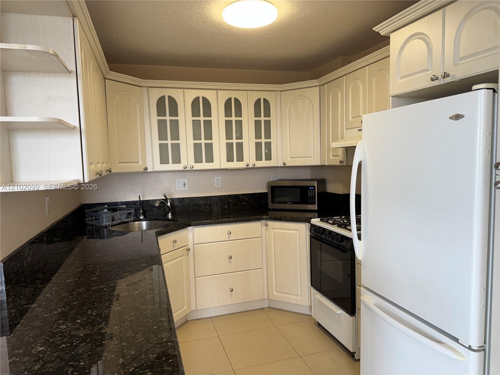 Kitchen featuring light-toned cabinetry with glass-panel upper doors, dark stone-finish countertops, tile flooring, and integrated white appliances