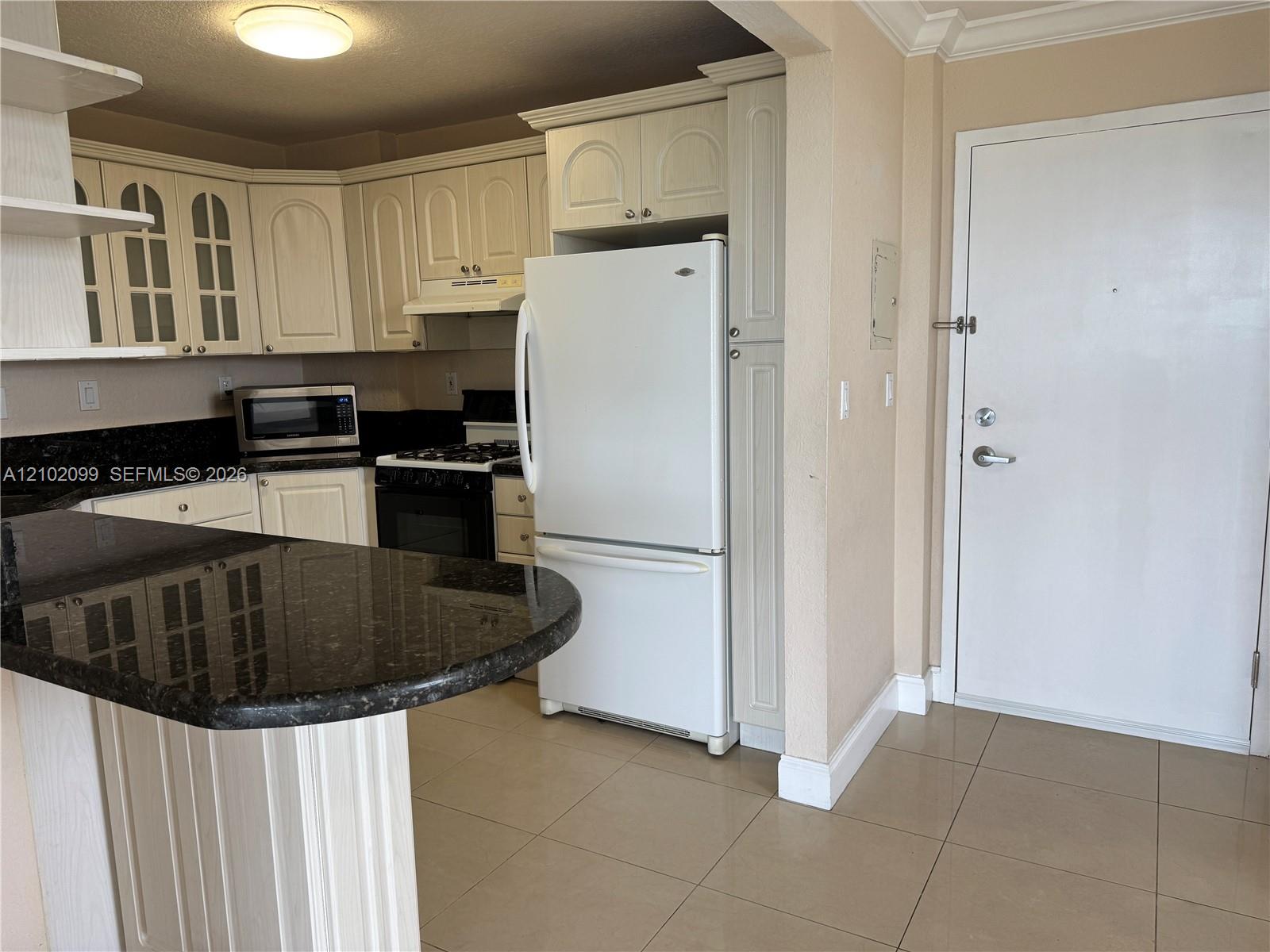 Kitchen featuring a peninsula with dark stone countertops, off-white cabinetry with glass-panel upper doors, tile flooring, and integrated white appliances