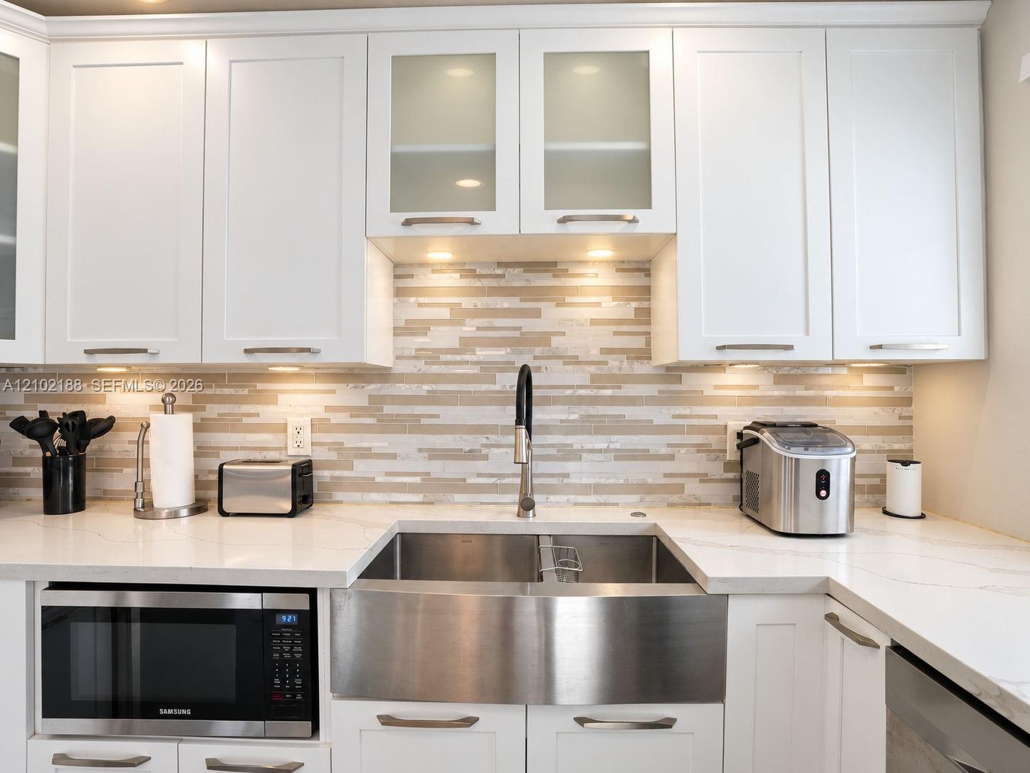 Kitchen featuring white shaker-style cabinetry with glass-front uppers, solid surface countertops, a horizontal mosaic tile backsplash, and an undermount stainless steel farmhouse sink with a high-arc pull-down faucet